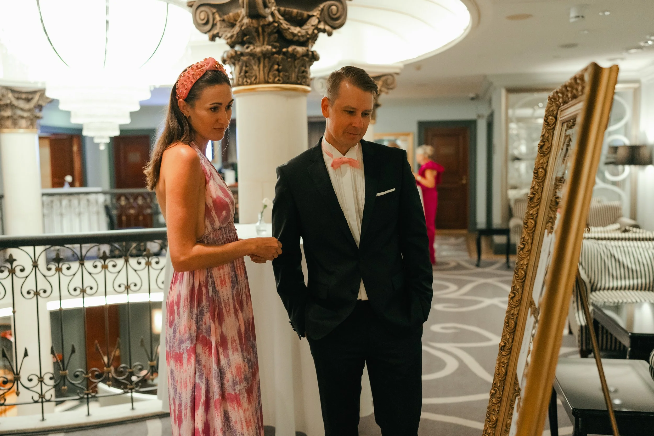 A man and woman dressed formally, standing in a luxurious interior with ornate pillars and chandeliers, looking at a framed display or mirror.