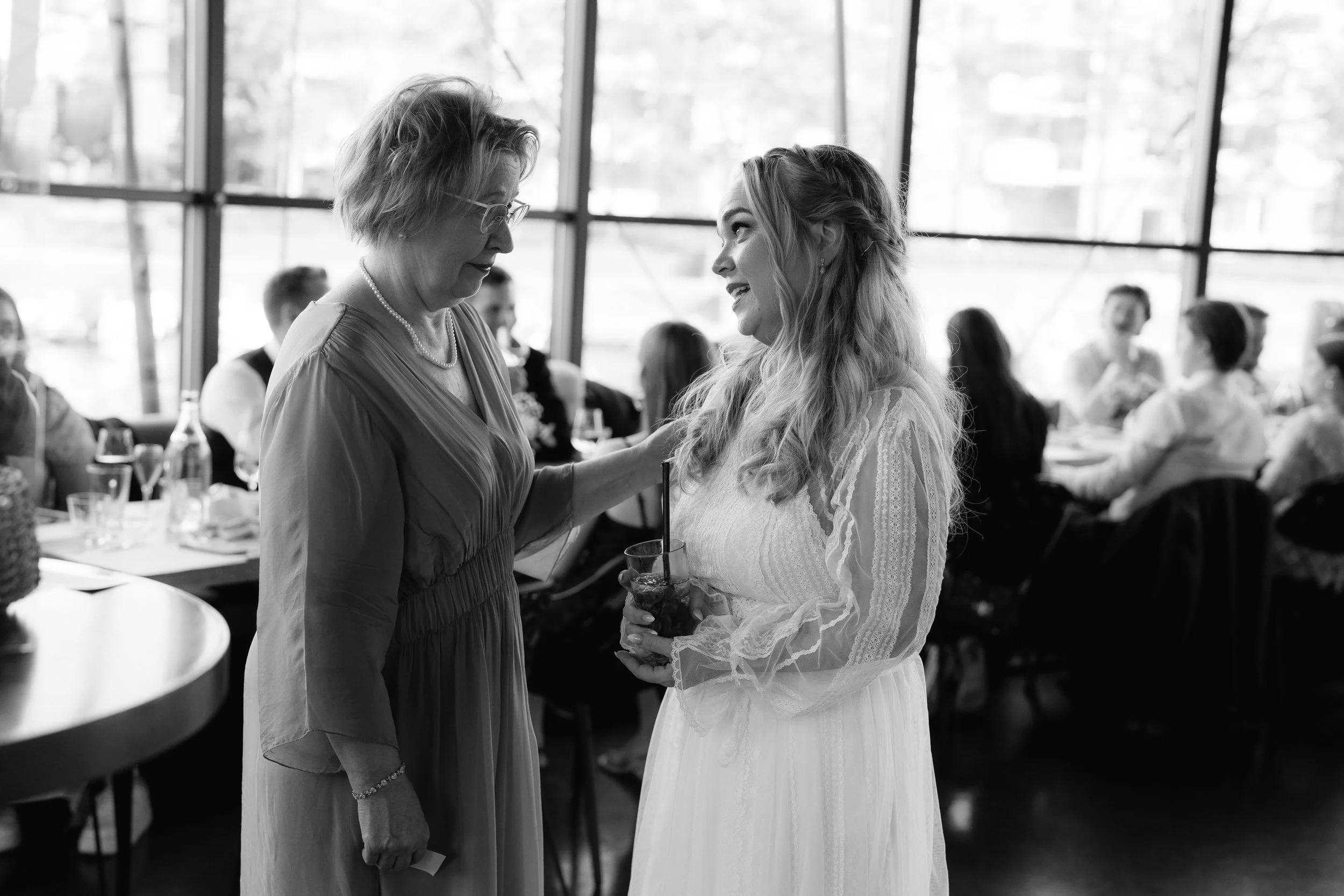 Black and white photo of two women talking at a formal event, with other guests seated in the background.
