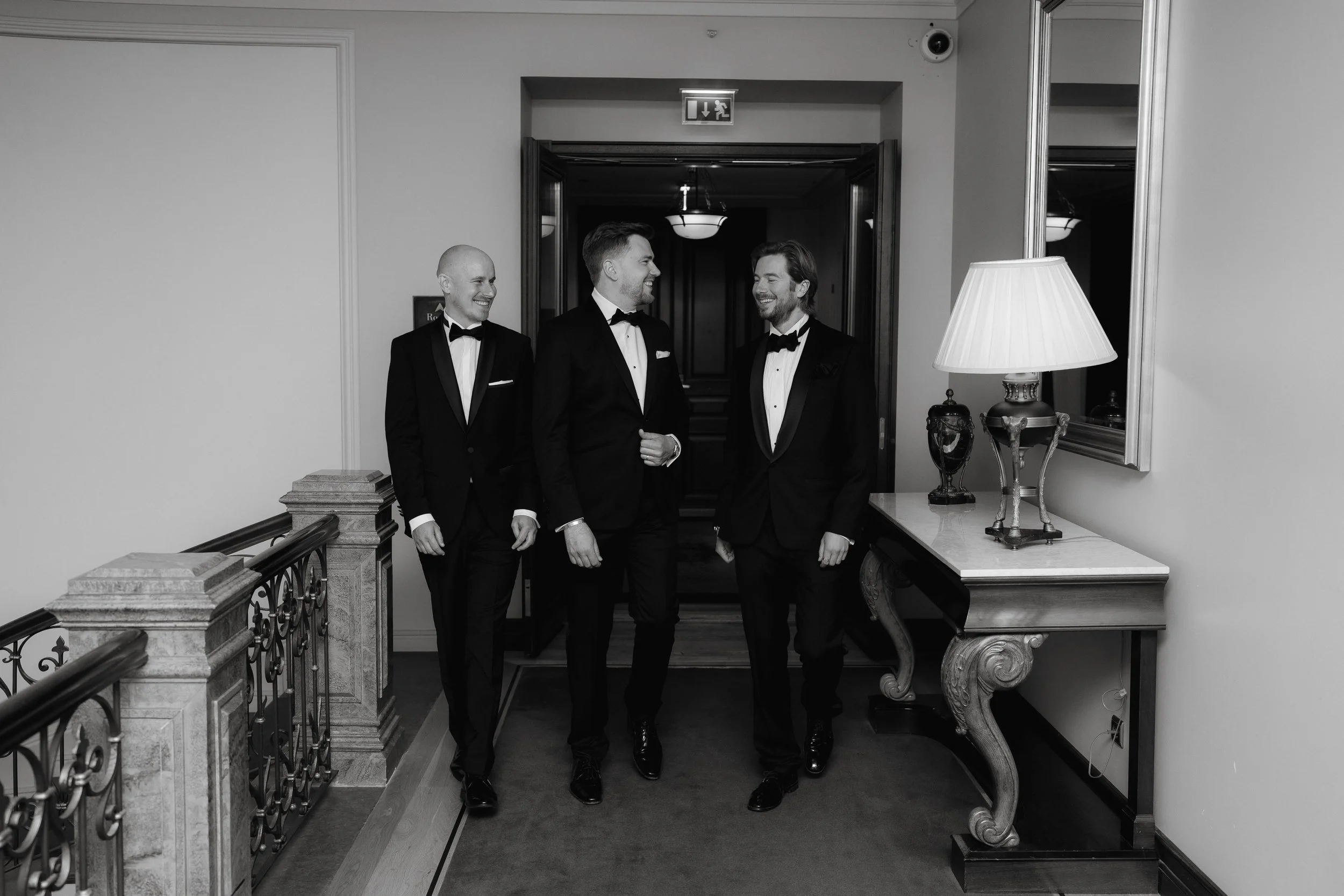 Three men in tuxedos walking indoors, black and white photo, elegant setting with a lamp on a side table.