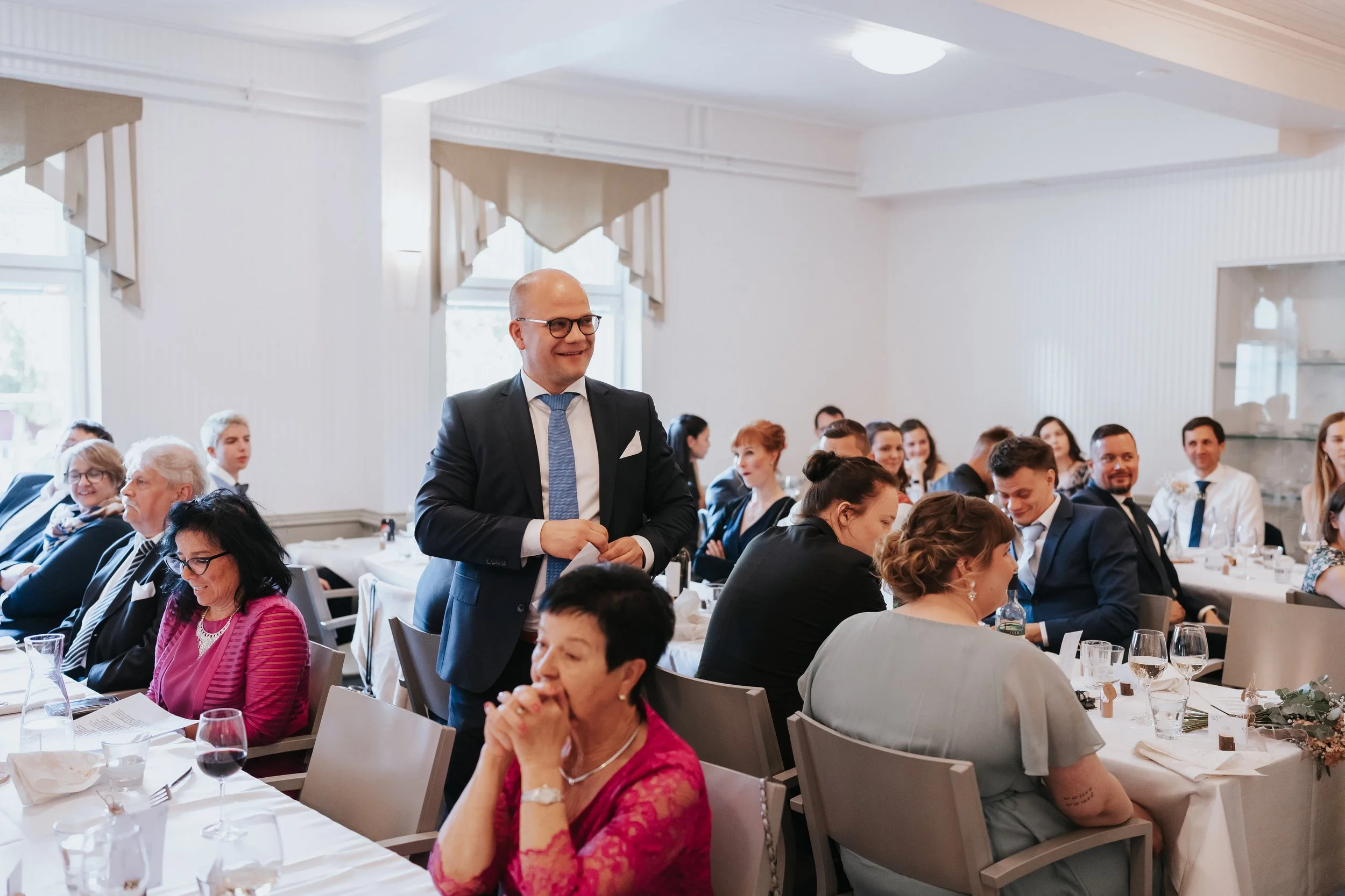 A gathering of people dressed in formal attire sitting at tables in a bright room, likely at a social event or reception.