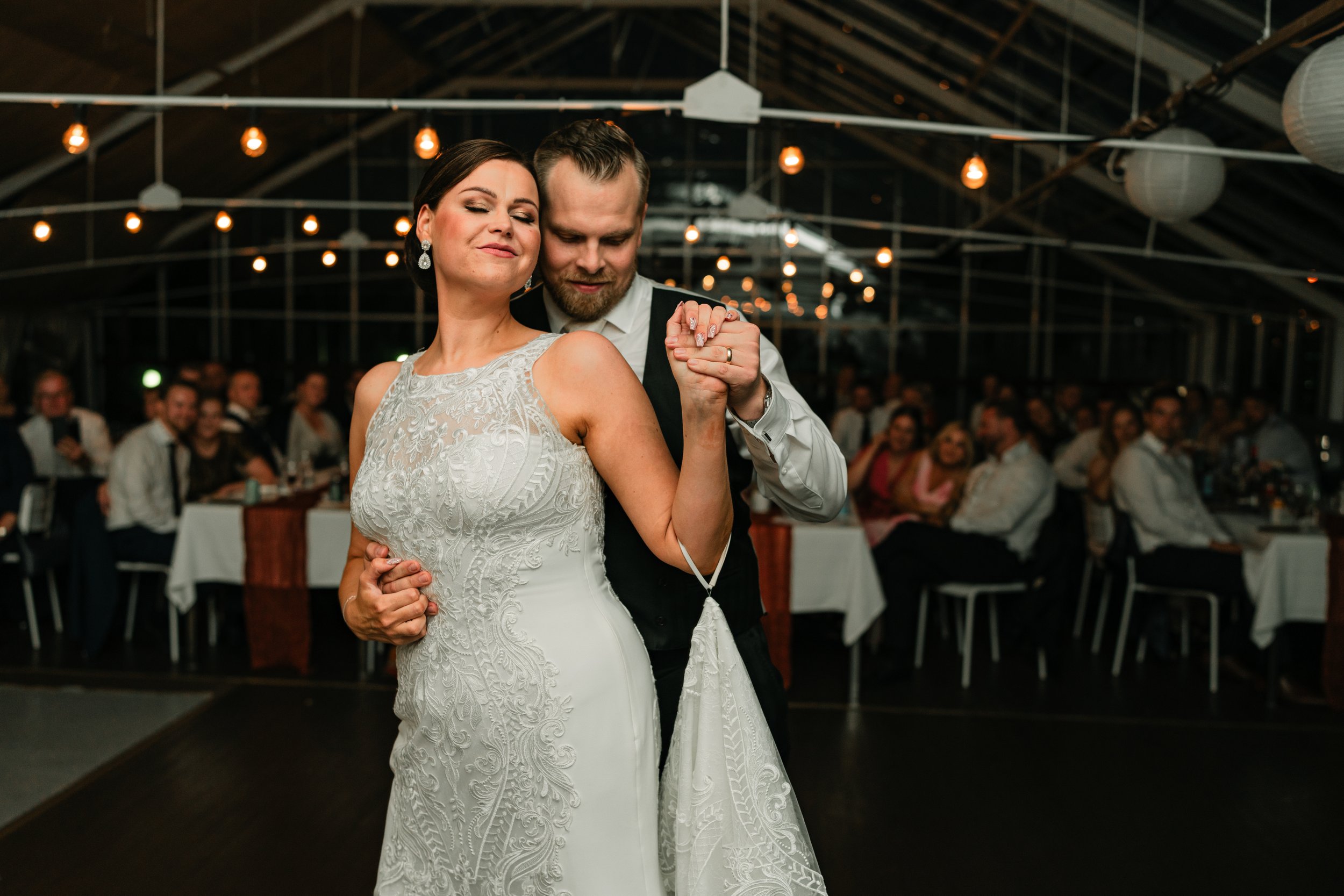 Couple dancing at a wedding reception, bride in a white dress, groom in a suit, with guests in the background.