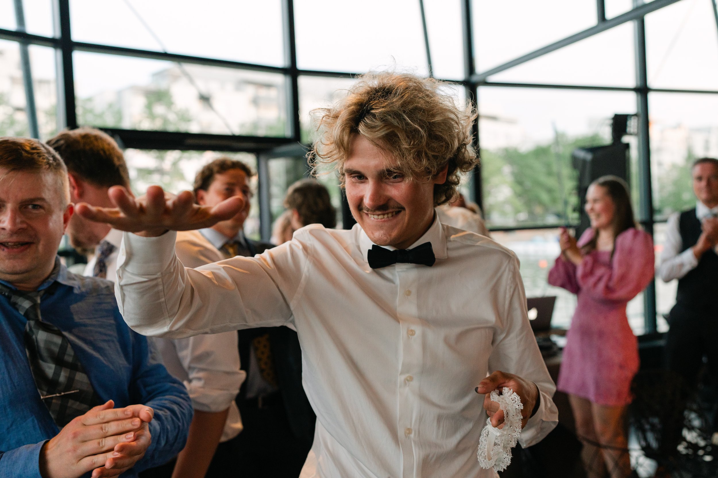 Man in white shirt and bow tie holding a garter surrounded by people at an event.