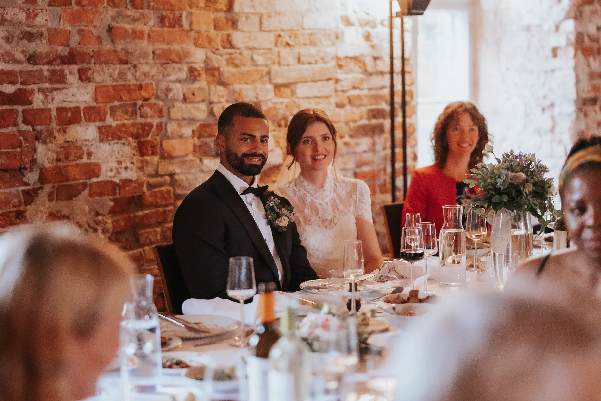 Wedding reception scene with a smiling bride and groom seated at a table, surrounded by guests in a rustic brick-walled room.