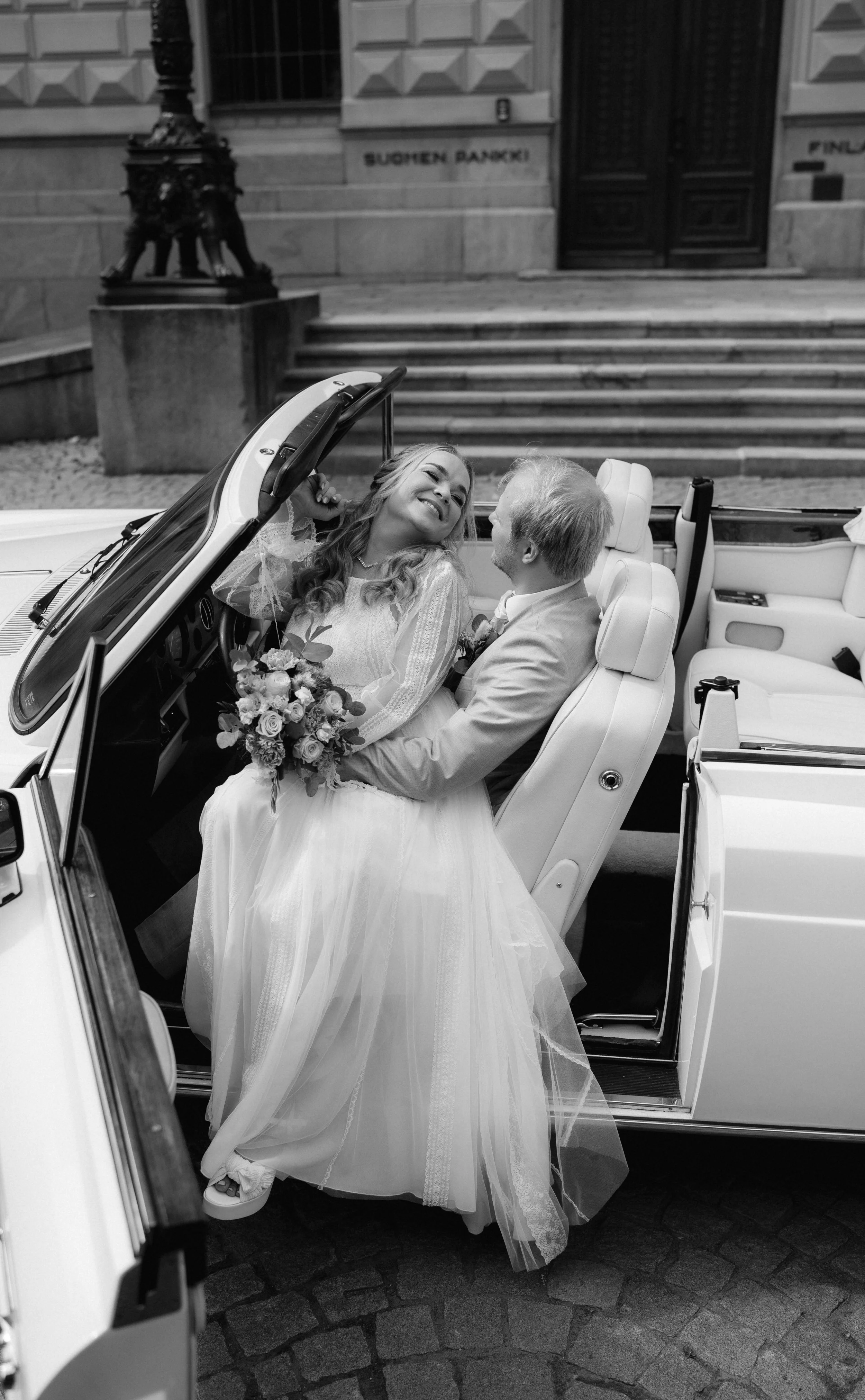 Bride and groom sitting in convertible car holding bouquet, black and white photo
