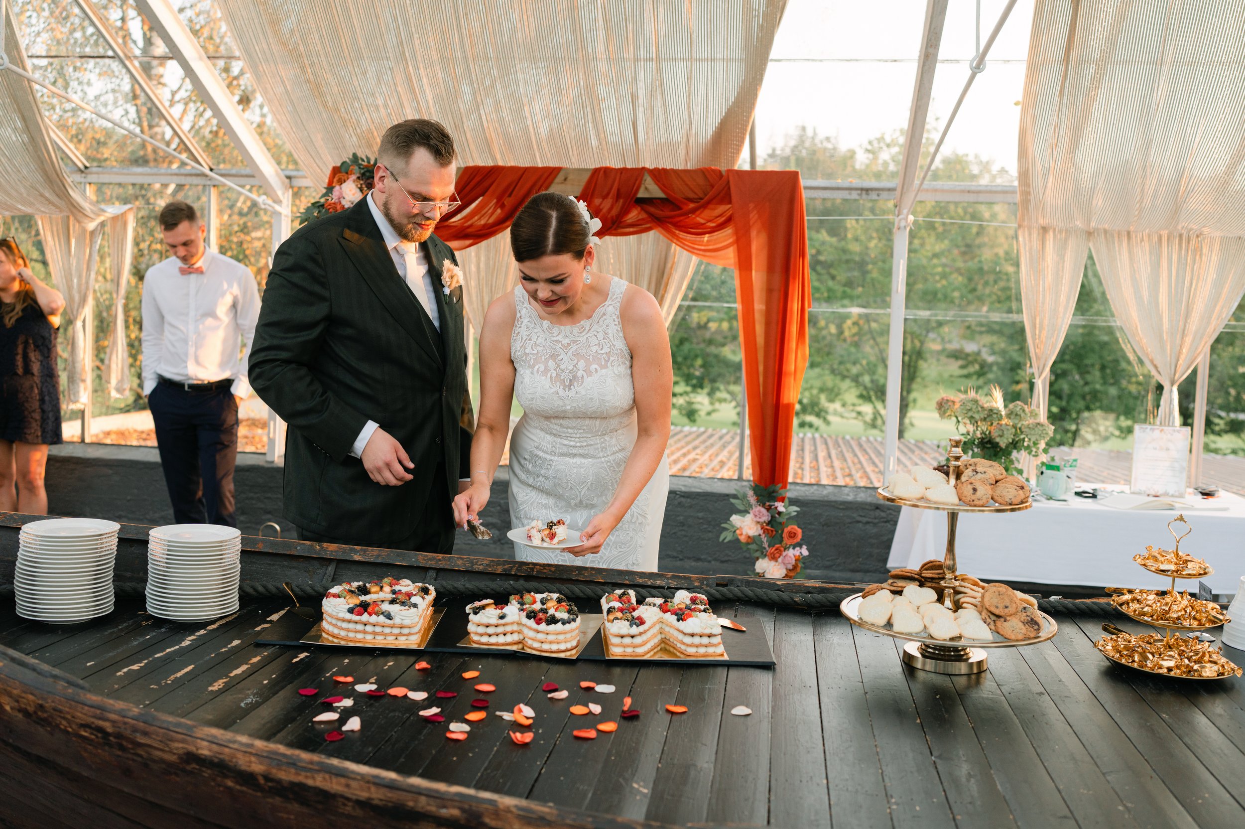 Wedding couple cutting cake at reception table with layered desserts, plates, and cookies displayed; decorative drapery and floral arrangements in background.