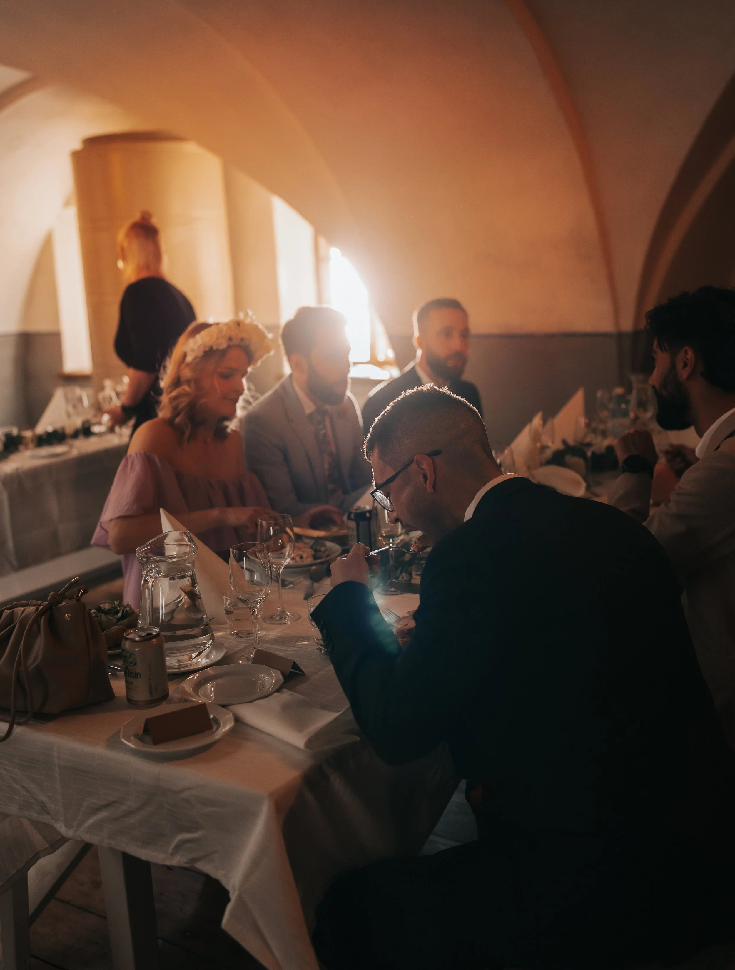 People sitting at a dinner table in a dimly lit room, engaged in conversation, with food and drinks on the table.