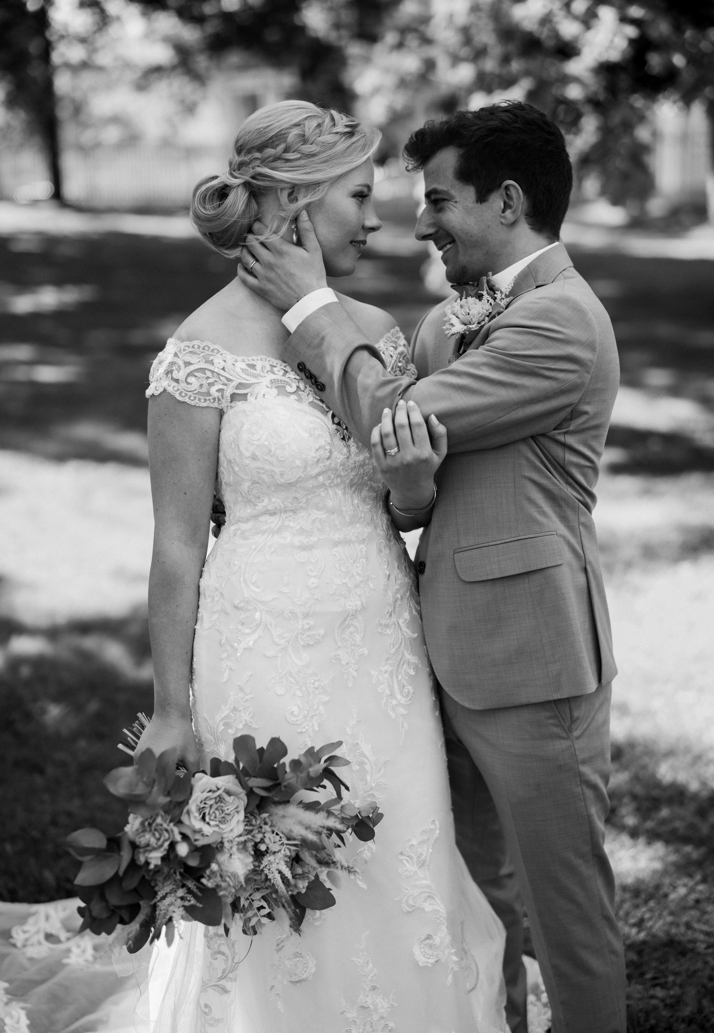 Bride and groom embracing outdoors, black and white photo, wedding dress with lace details, holding bouquet, romantic moment.