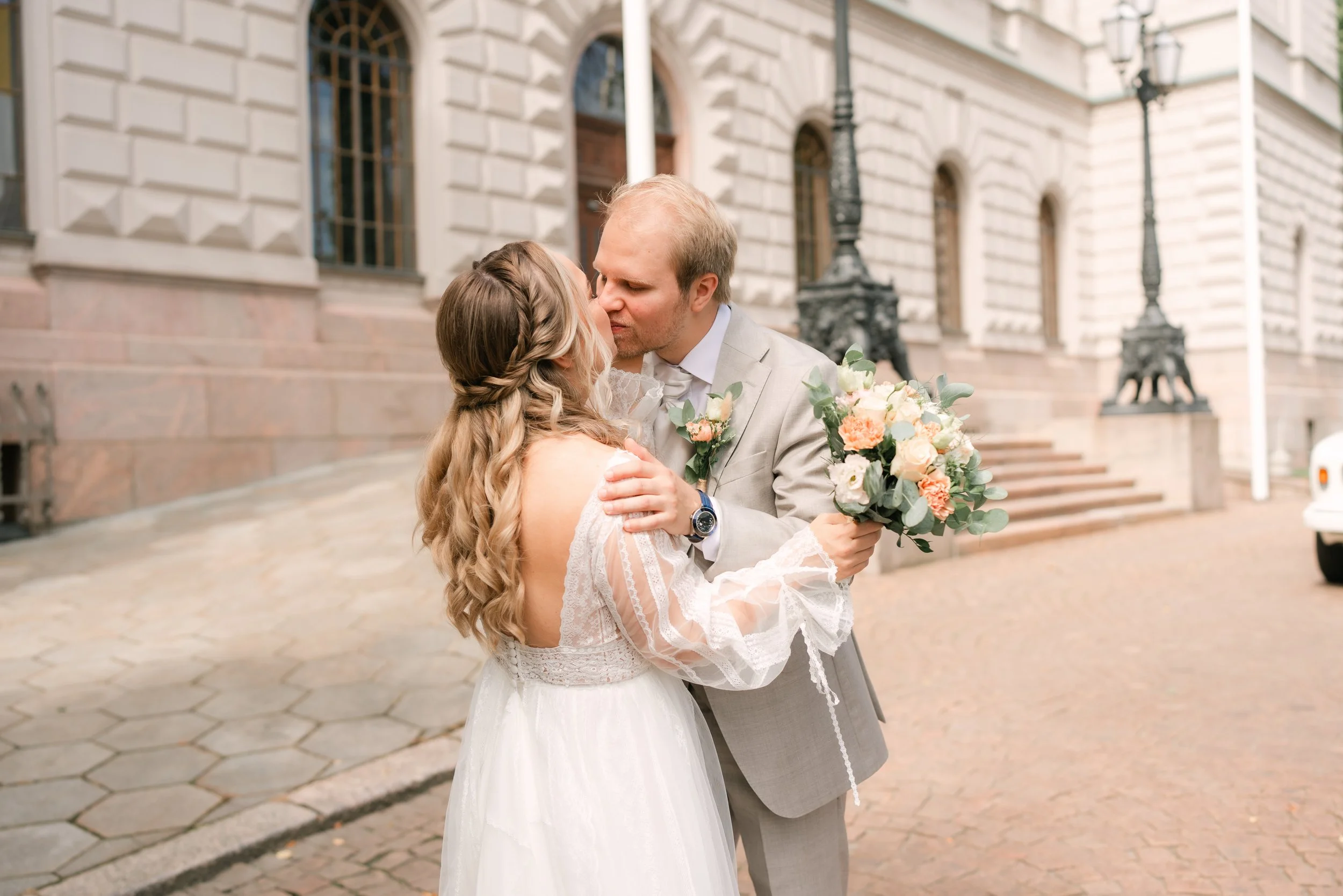Bride and groom kissing on street, bride holding bouquet, historic building in background.