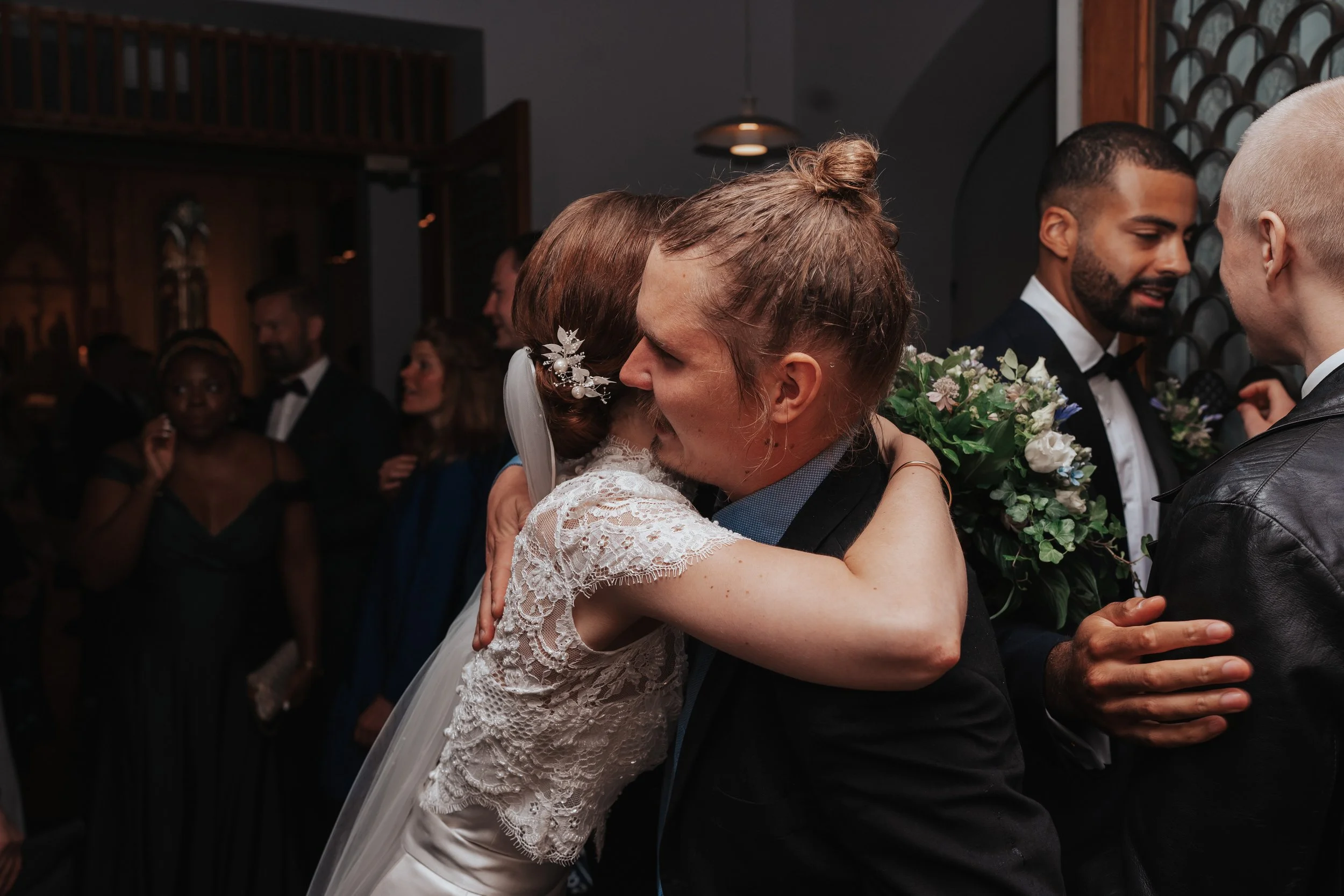 Wedding scene with bride hugging a guest in formal attire, surrounded by people socializing.