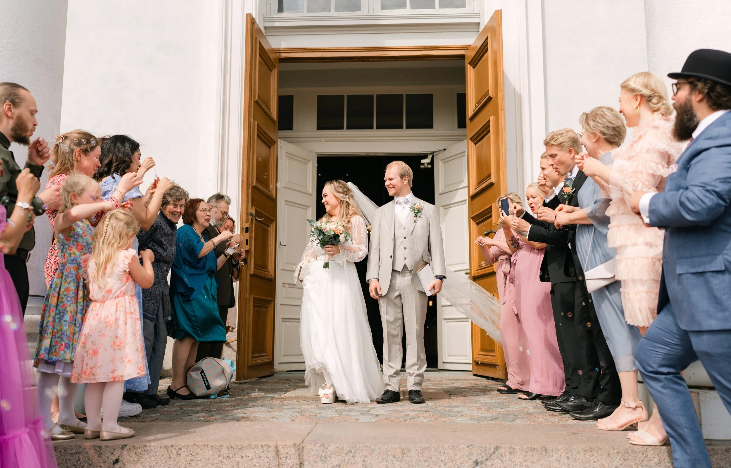 Bride and groom exiting a building, surrounded by guests celebrating, wearing formal attire, with open wooden doors.