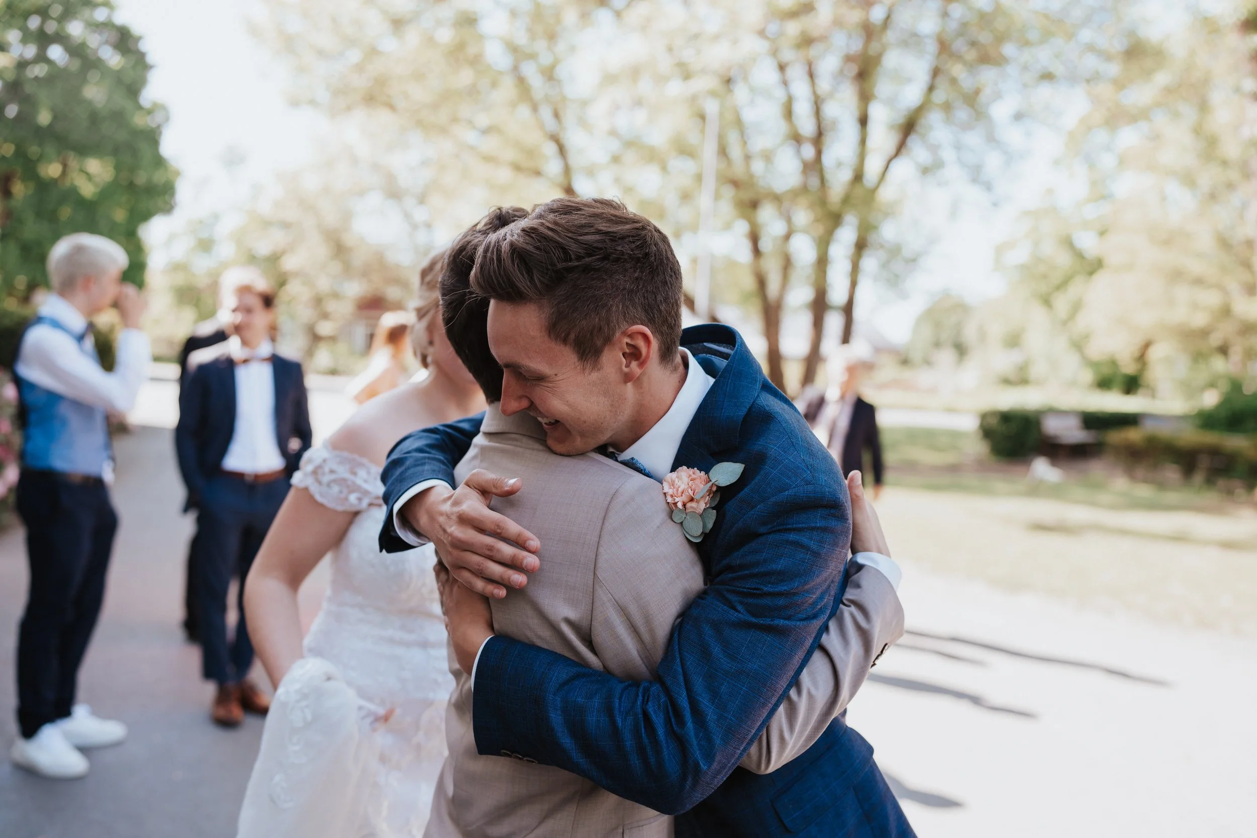 Wedding guests hugging outdoors with others in suits and dresses in background.