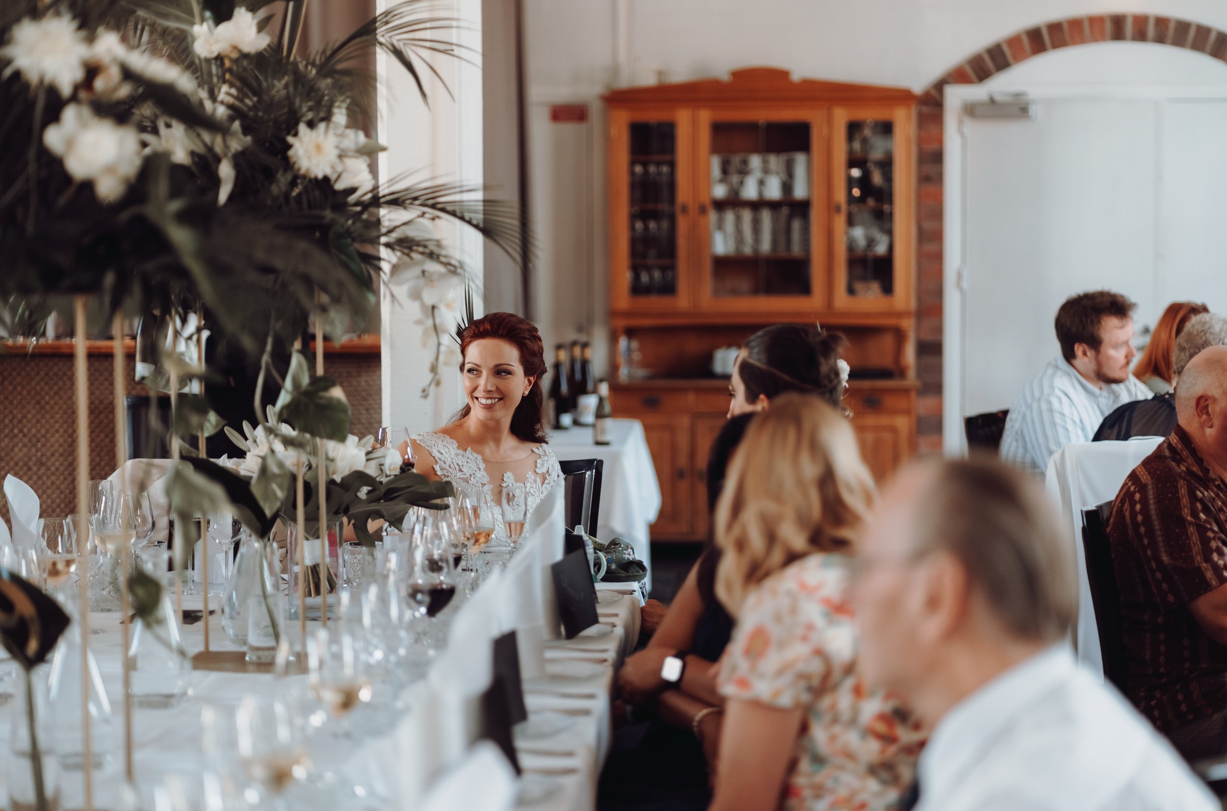 Indoor event with people seated at a decorated table with floral arrangements and a wooden cabinet in the background.