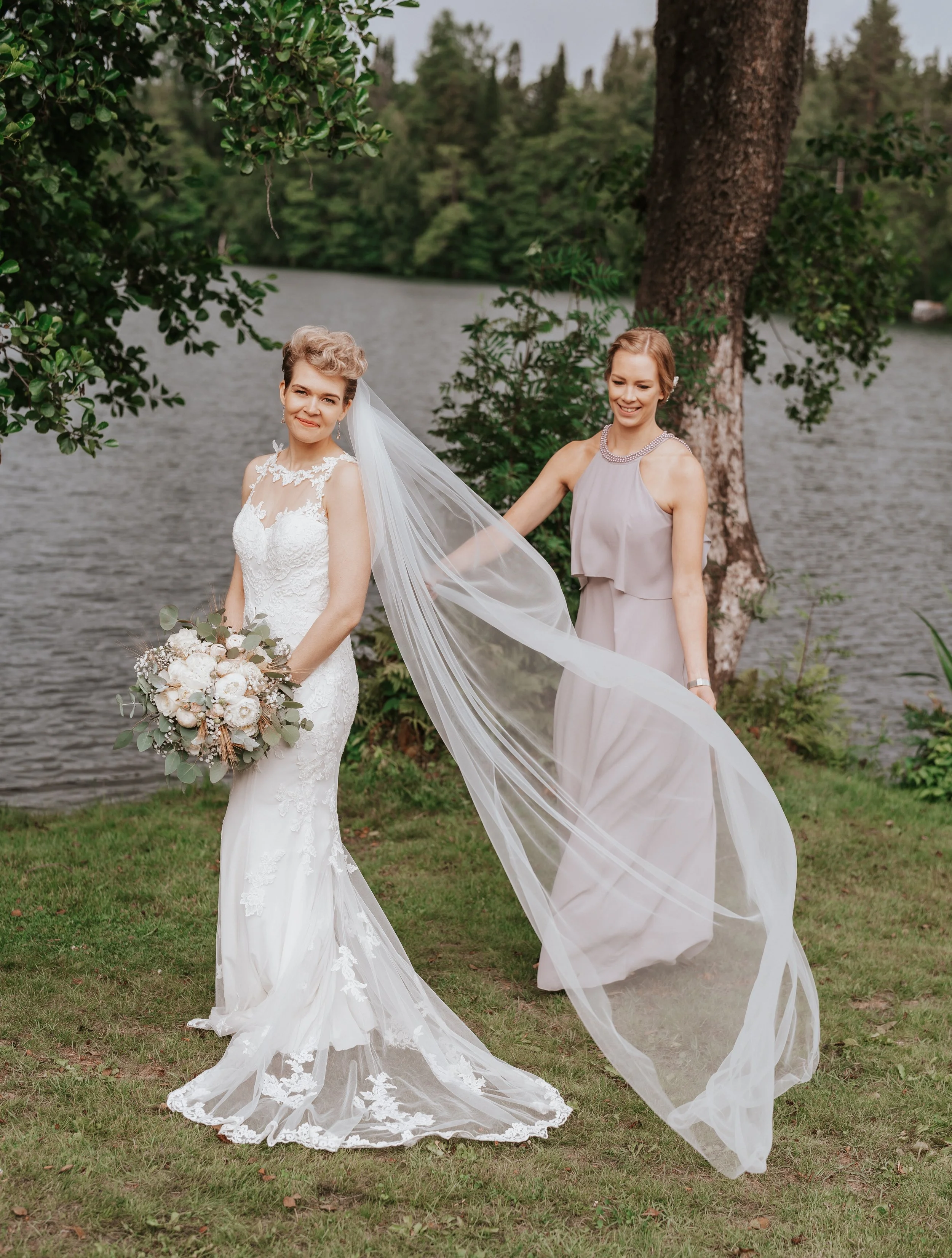 Bride in lace wedding dress holding bouquet, with bridesmaid adjusting long veil, standing by a lake, surrounded by trees.