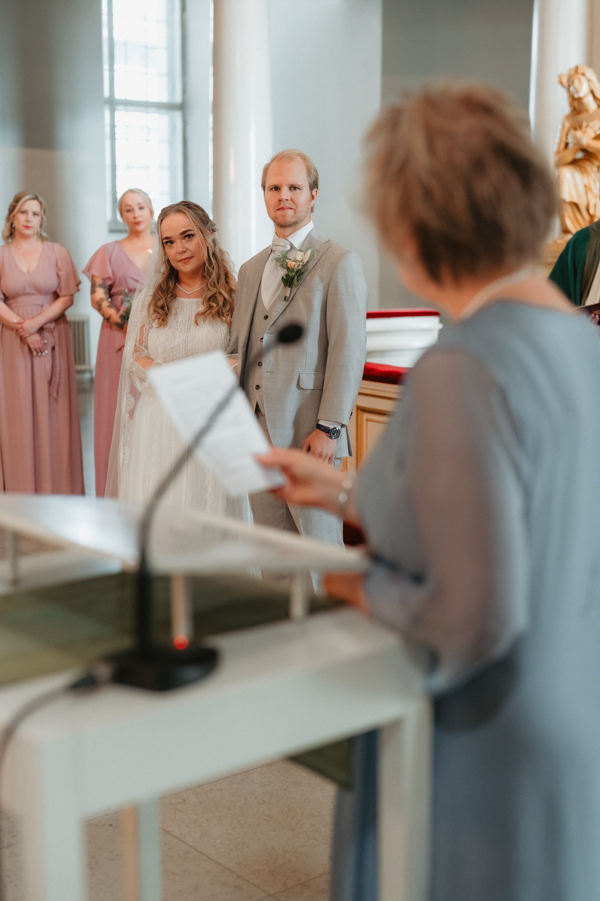 Bride and groom standing at a wedding ceremony with an officiant speaking at a podium, bridesmaids in the background wearing pink dresses.