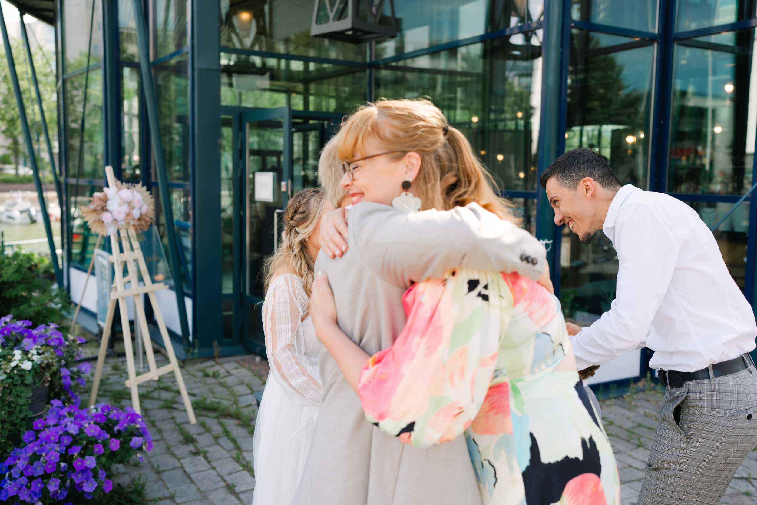 Four people outdoors, two women hugging, man smiling, near a glass building with flowers in foreground.