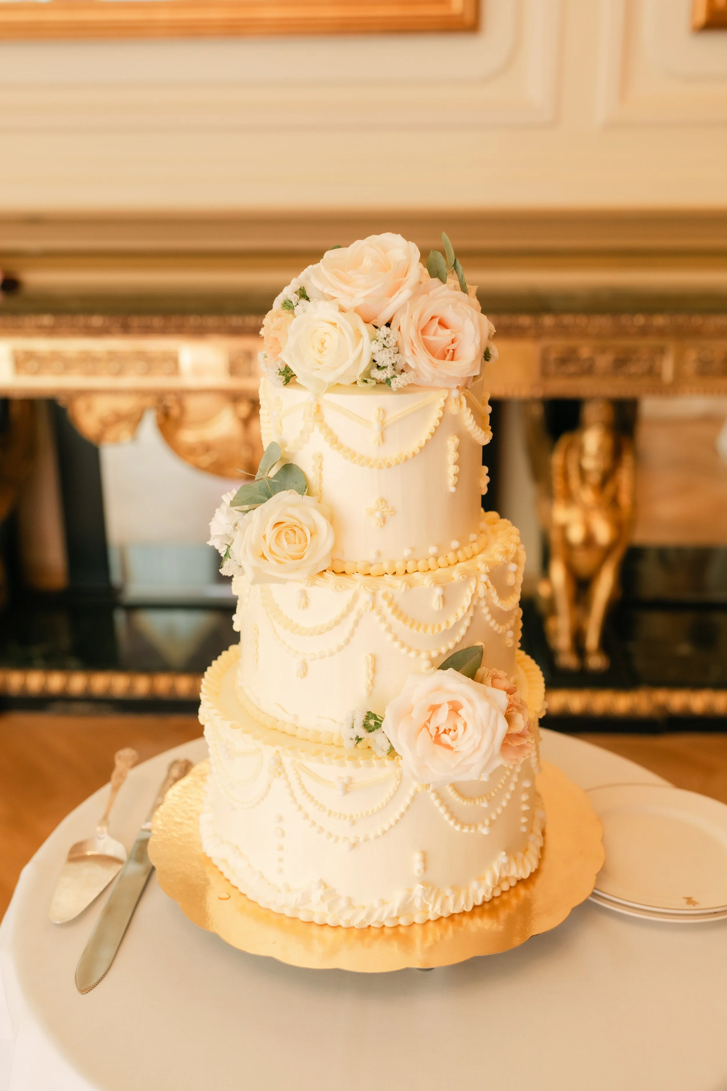 Three-tier wedding cake with white icing and pearl decorations, adorned with peach and white roses, on a table with a knife and serving fork.