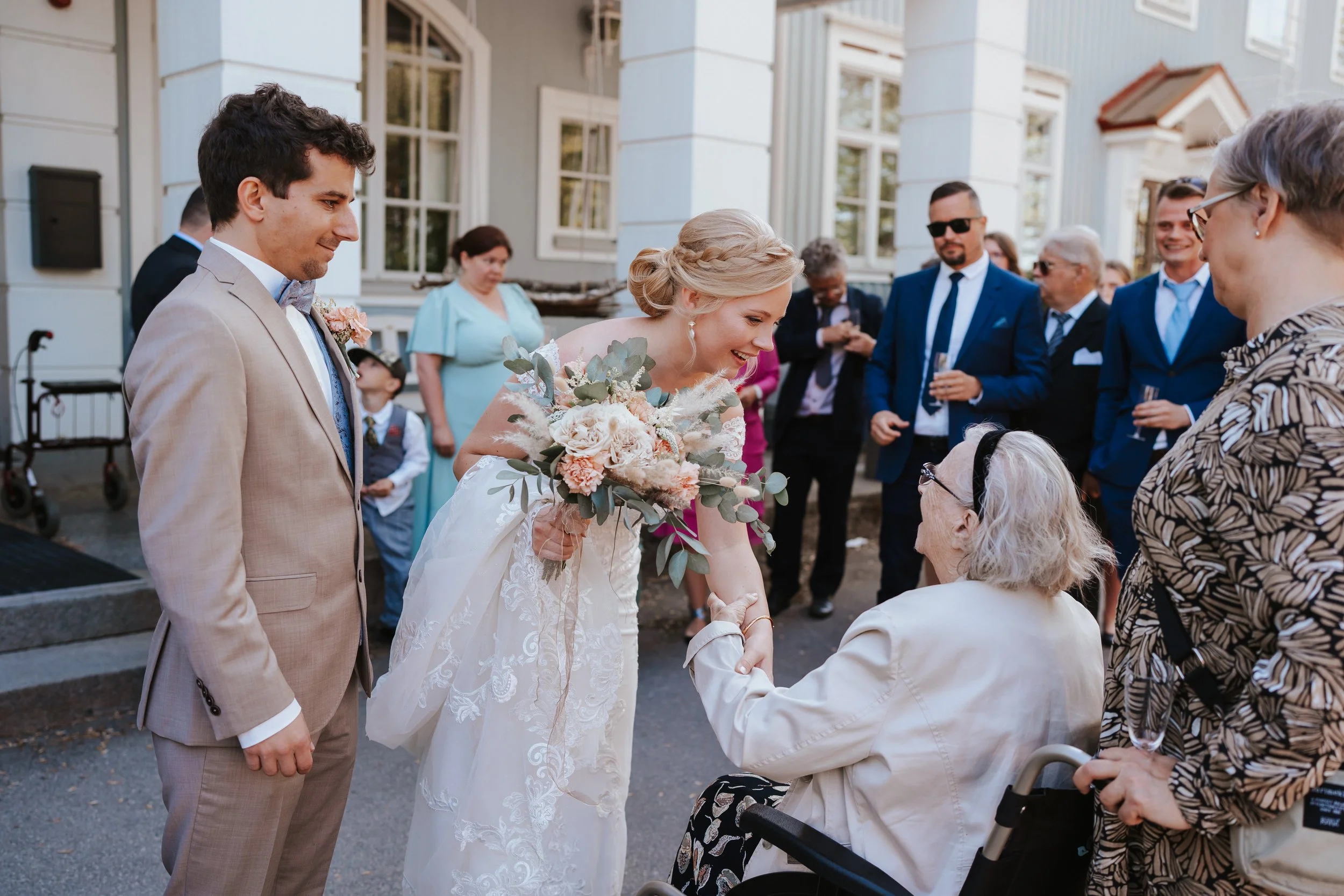Wedding scene with bride and groom greeting elderly woman in wheelchair, surrounded by elegantly dressed guests.