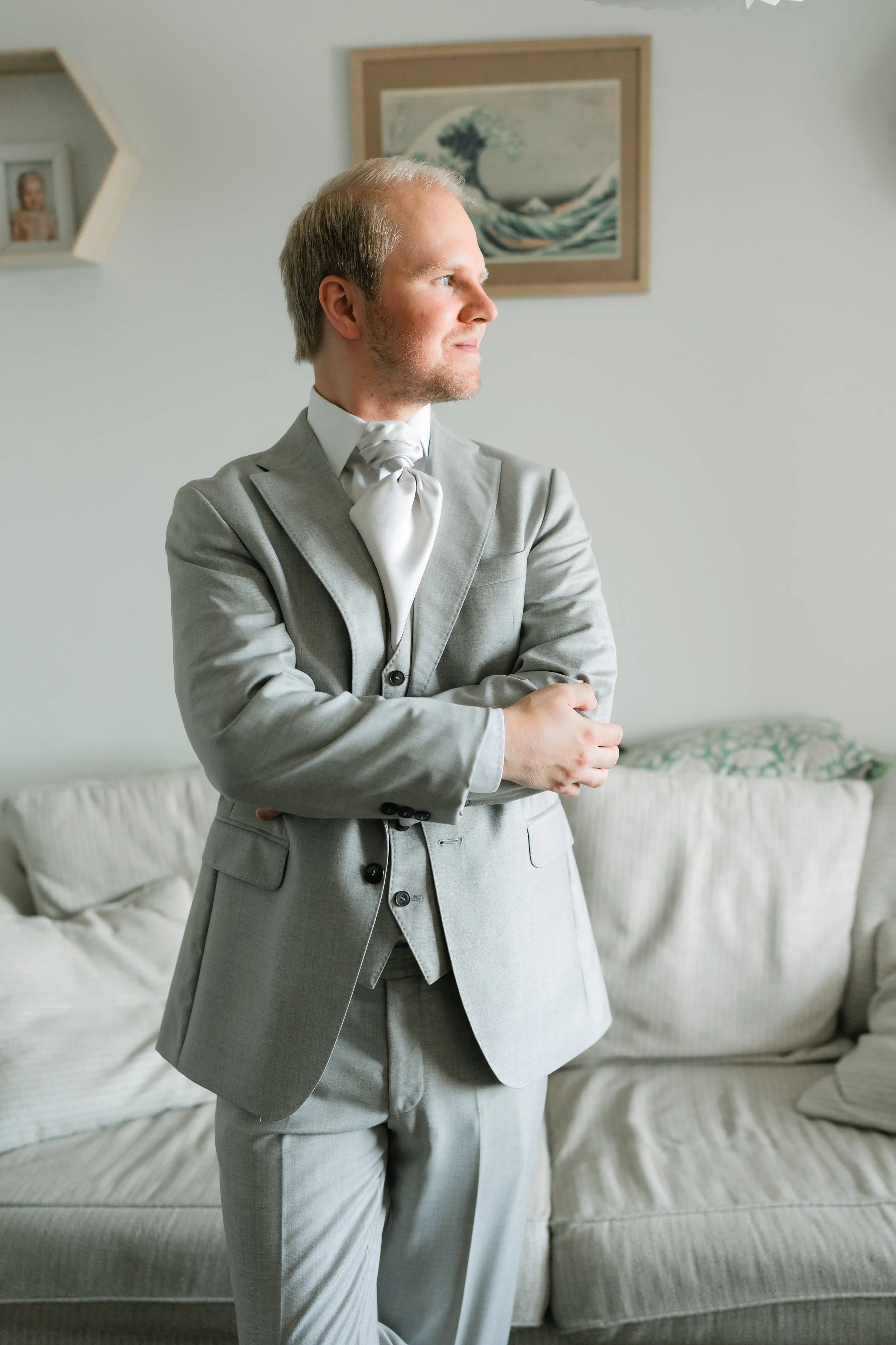 Man in a light gray suit with a white cravat standing in a living room.