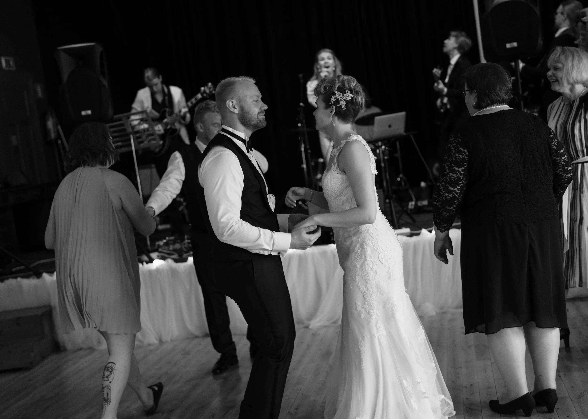 Black and white photo of a wedding reception with a bride in a white dress dancing, accompanied by guests and live musicians in the background.