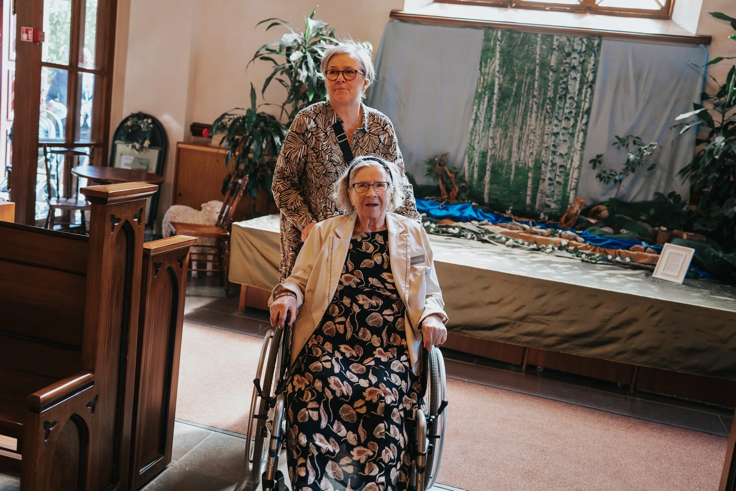 Two elderly women indoors, one in a wheelchair and the other standing behind, with a nature-themed display in the background.
