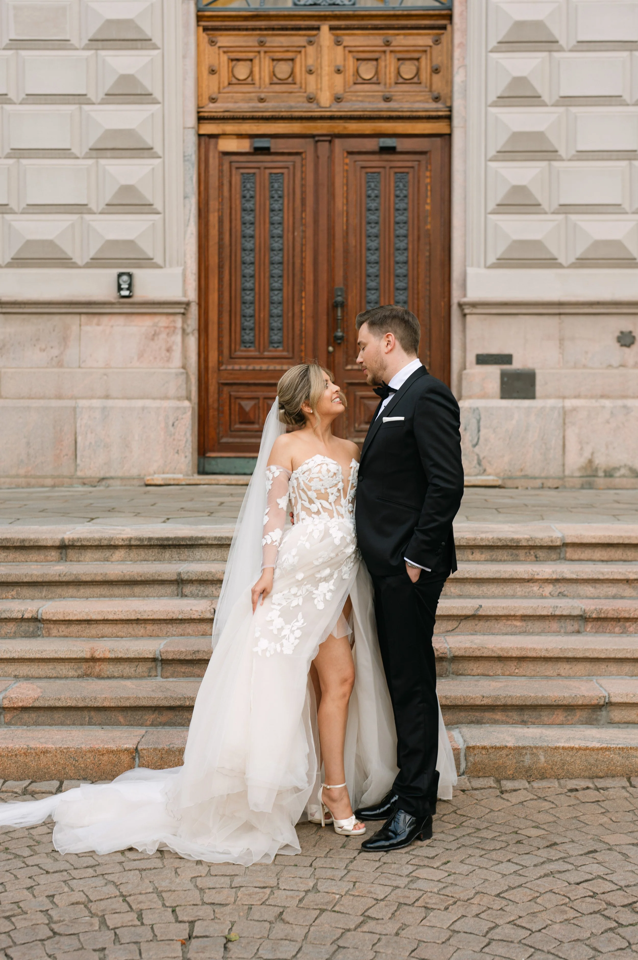 Bride and groom on wedding day, standing on stone steps in front of ornate wooden door, bride in lace gown, groom in black suit.