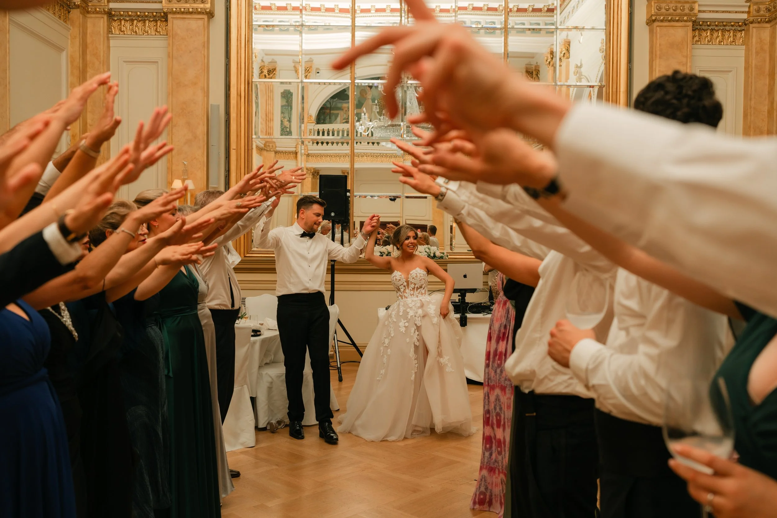 Bride and groom holding hands at a wedding reception with guests forming an arch of raised arms around them in an elegant venue.