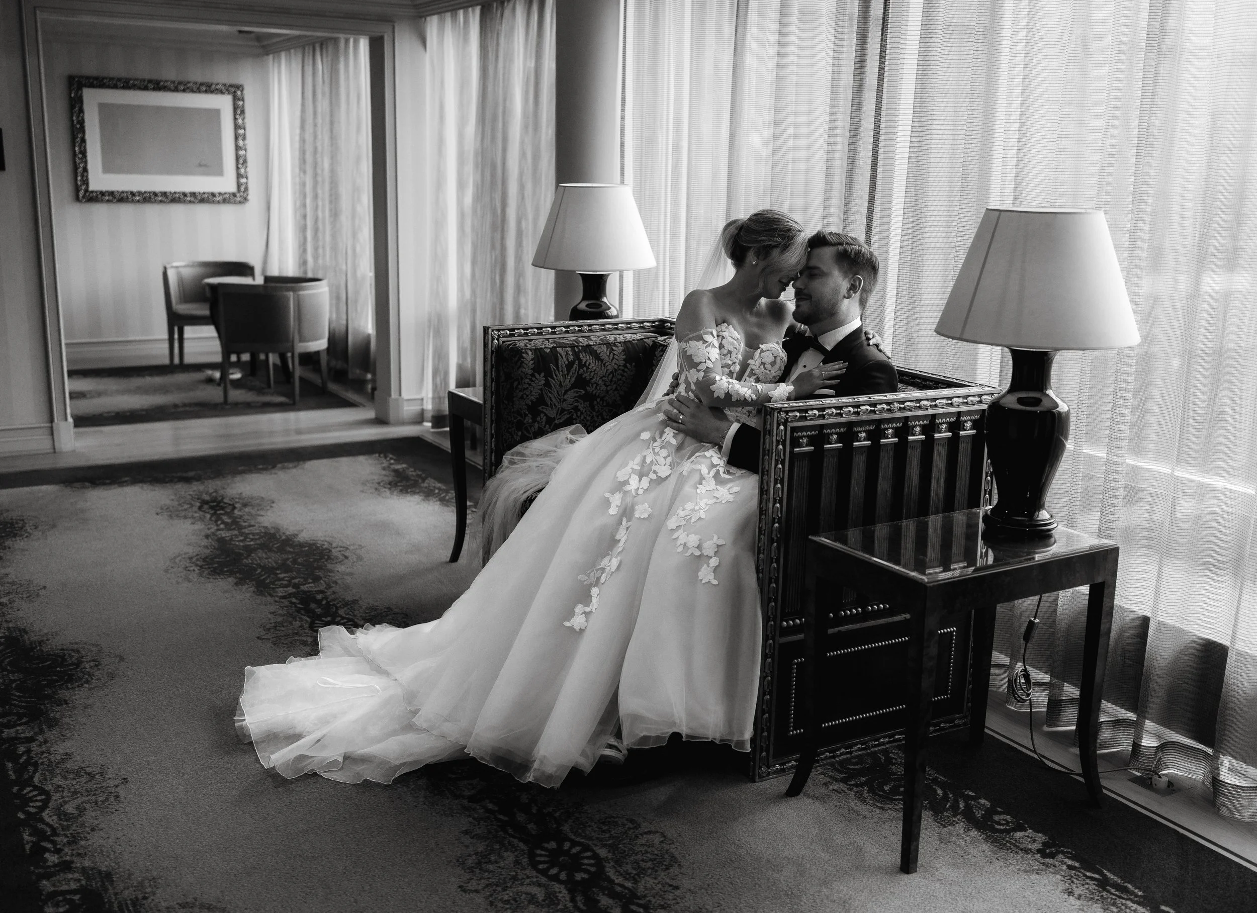 Black and white photo of a bride in an elaborate gown sitting closely with a groom in a suit on a decorative bench in a well-lit room.