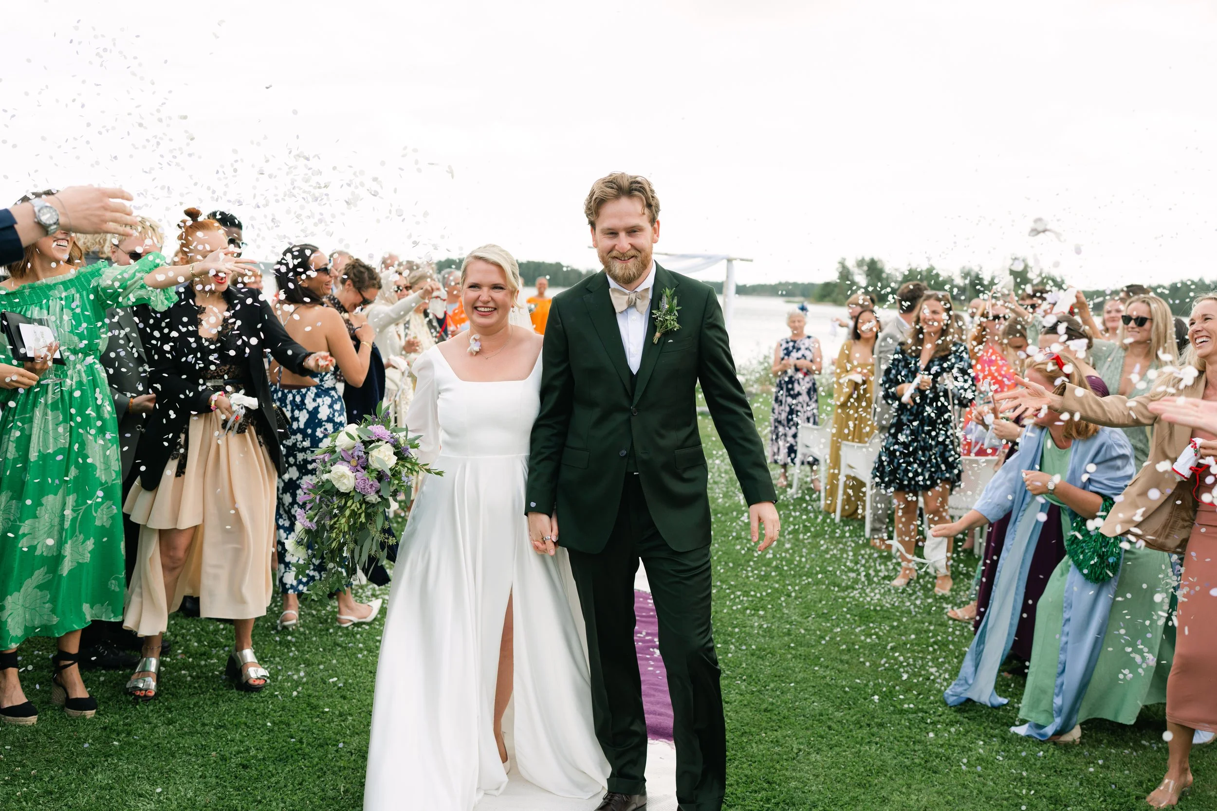 Bride and groom walking down the aisle with guests celebrating and throwing confetti near a lakeside outdoor wedding ceremony.