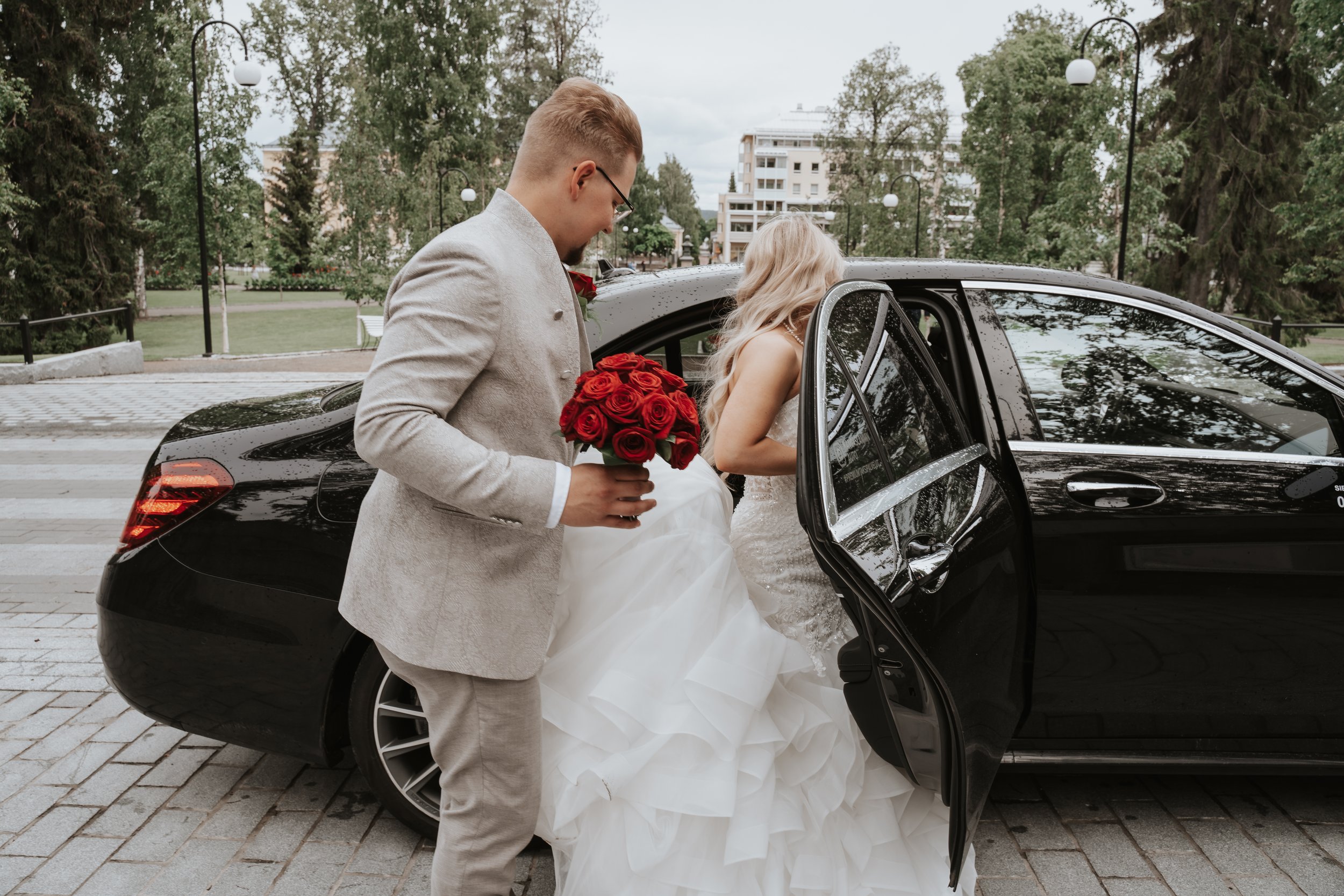 Bride getting into a black car while groom holds red roses