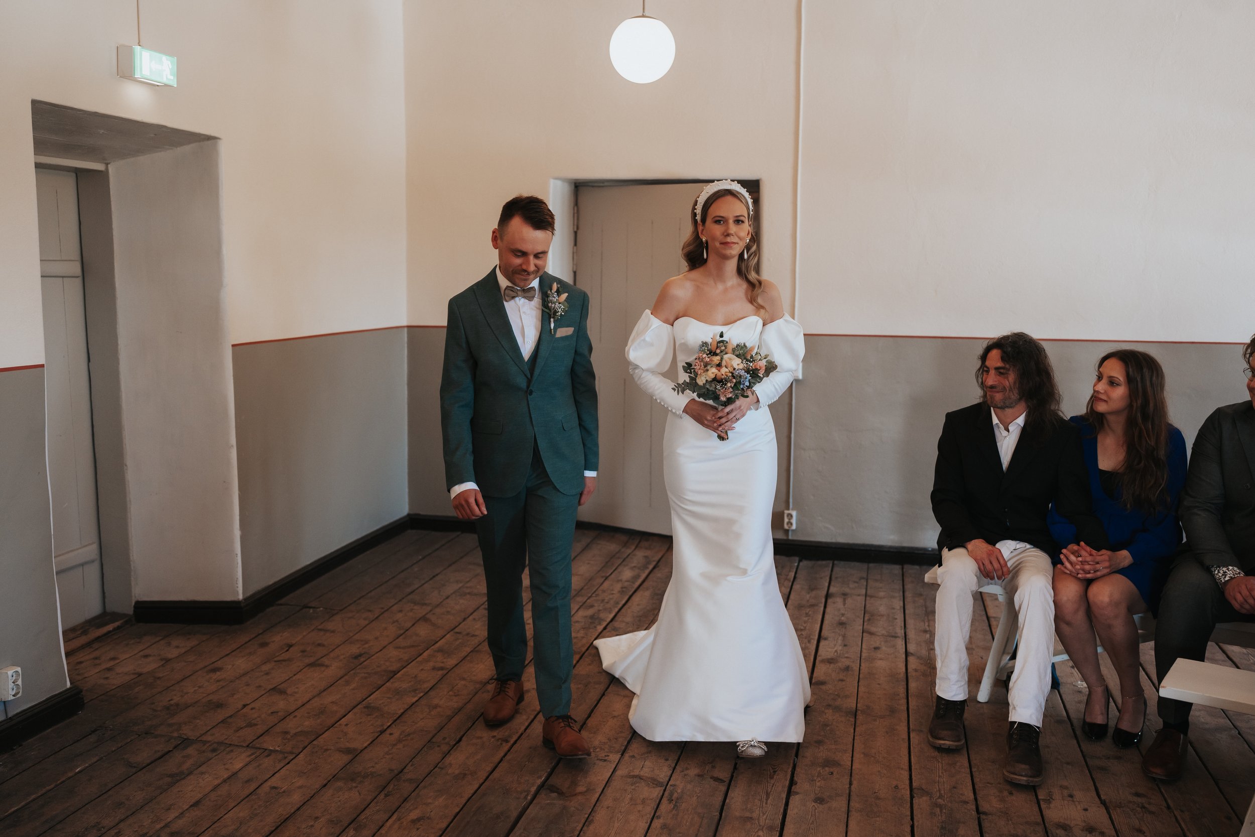 Bride and groom walking indoors at wedding ceremony, guests seated, wooden floor, minimalist setting.