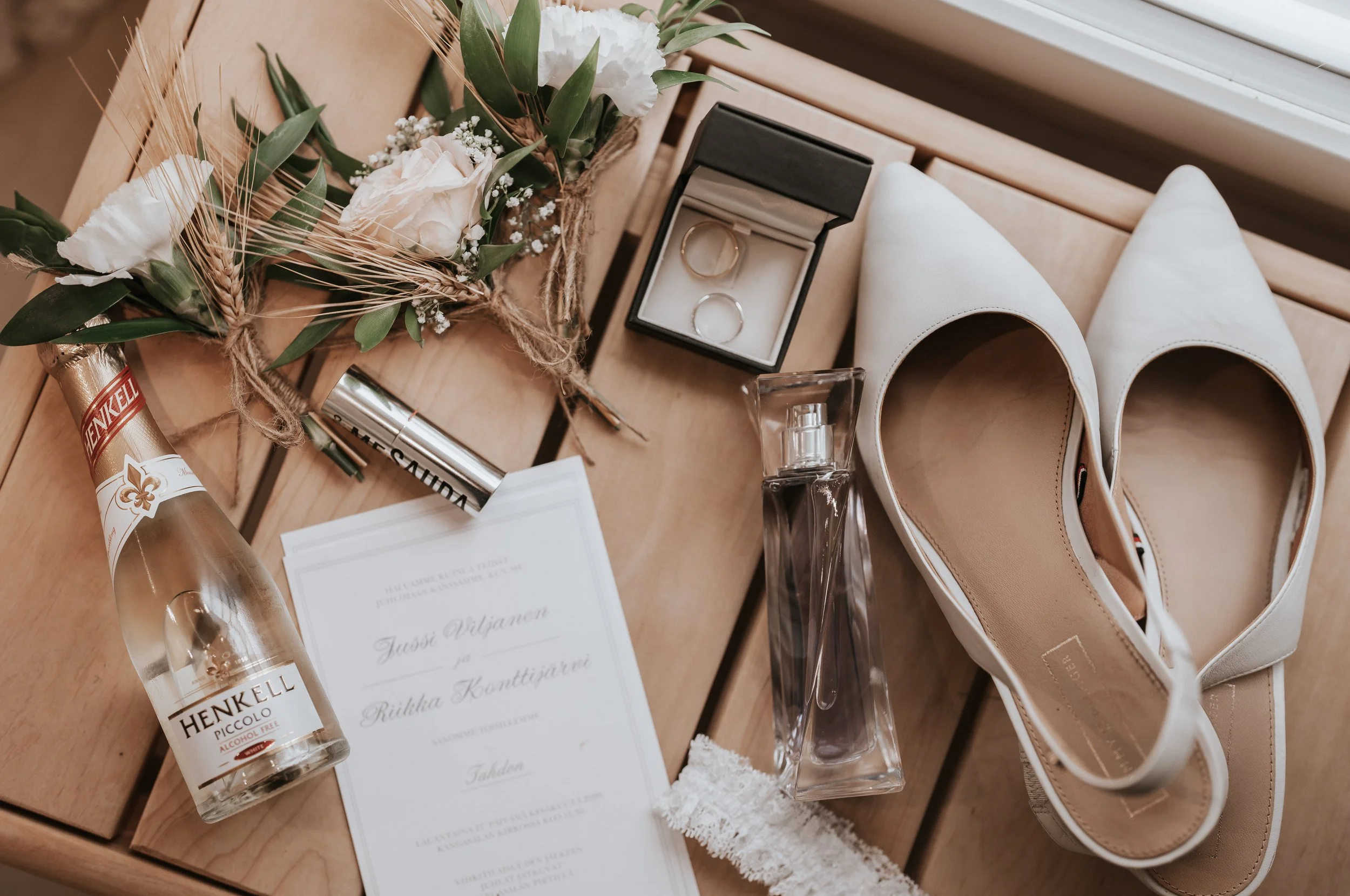 Wedding flat lay with white shoes, invitation, champagne, rings, bouquet, mascara, and perfume on wood surface.
