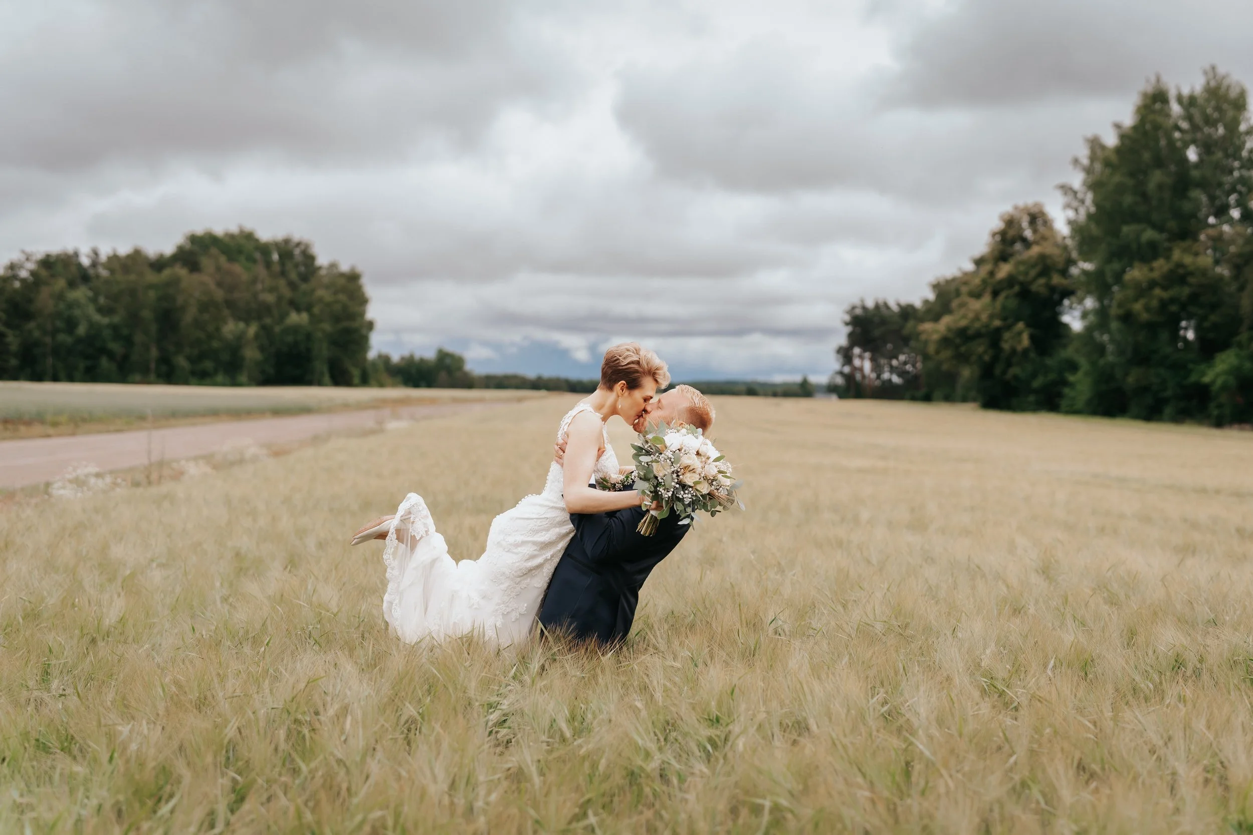 Bride and groom in wedding attire embracing in a field with cloudy sky background.