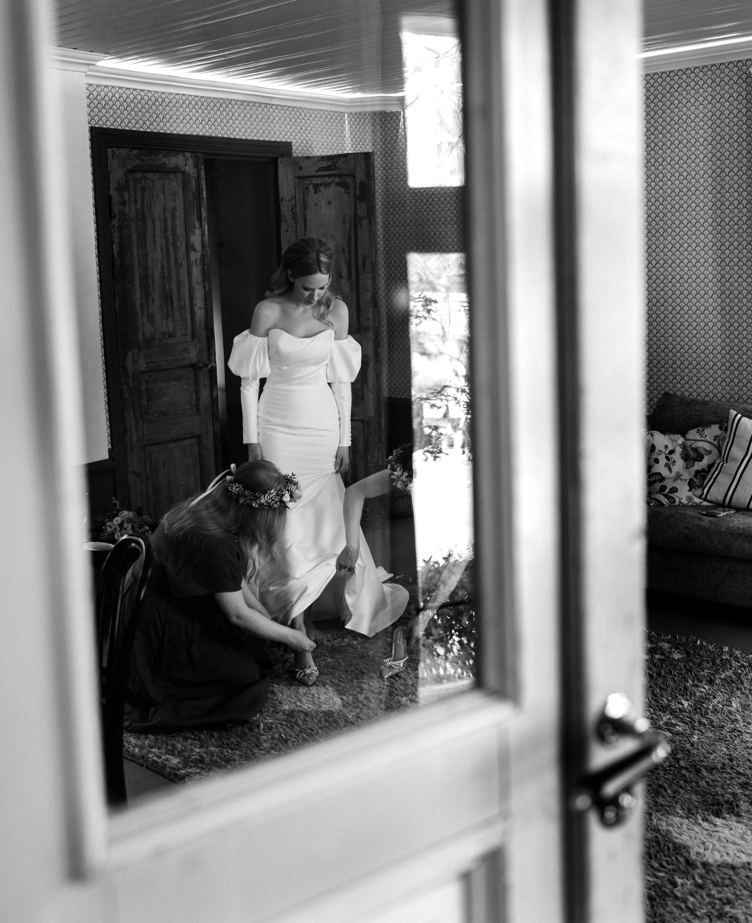 Black and white photo of a bride in an off-shoulder dress, bridesmaid helping with her gown, viewed through a glass door.