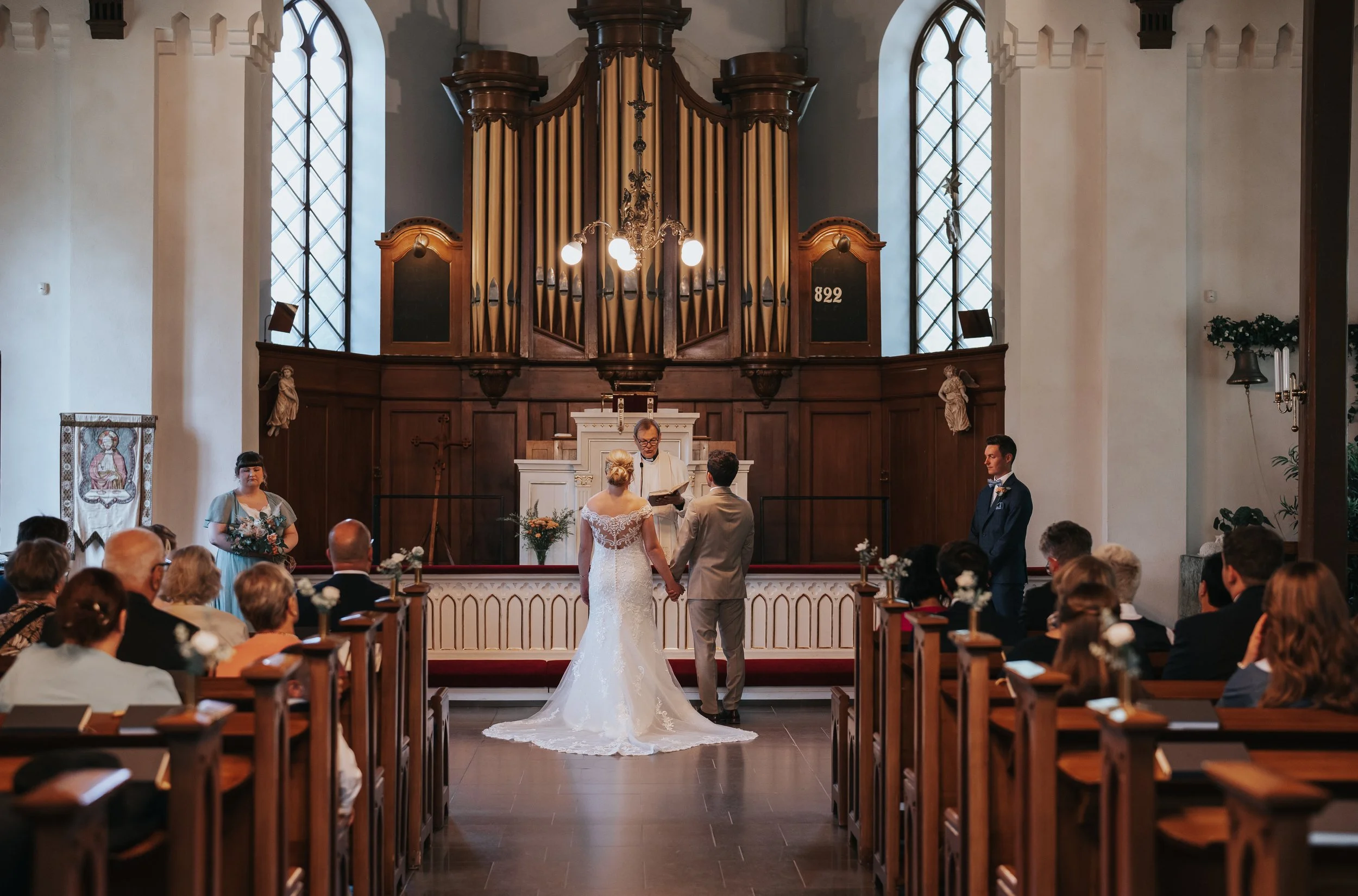 Wedding ceremony in a church with bride and groom holding hands at the altar, officiant conducting the service, guests seated, and large organ in the background.