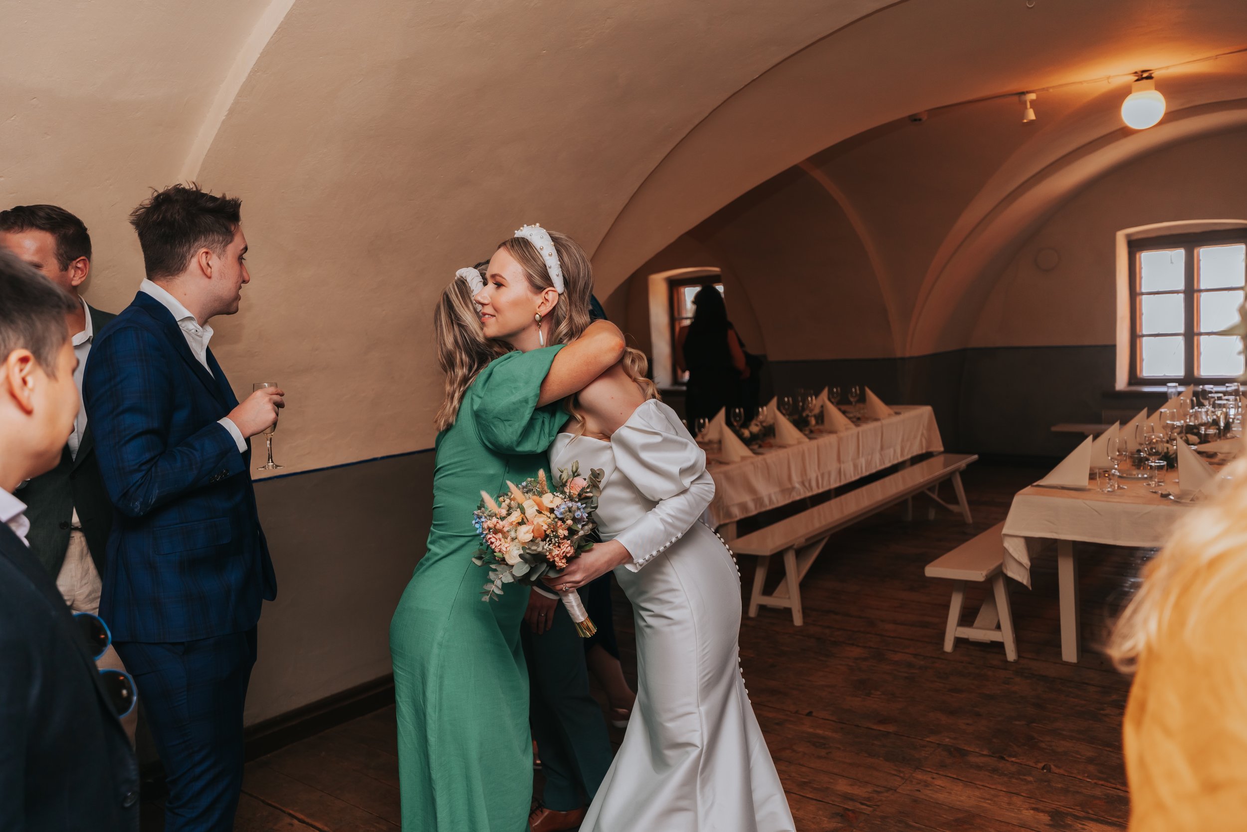 Bride hugging guest in green dress at wedding, holding bouquet, people in formal attire, decorated reception room with banquet tables.