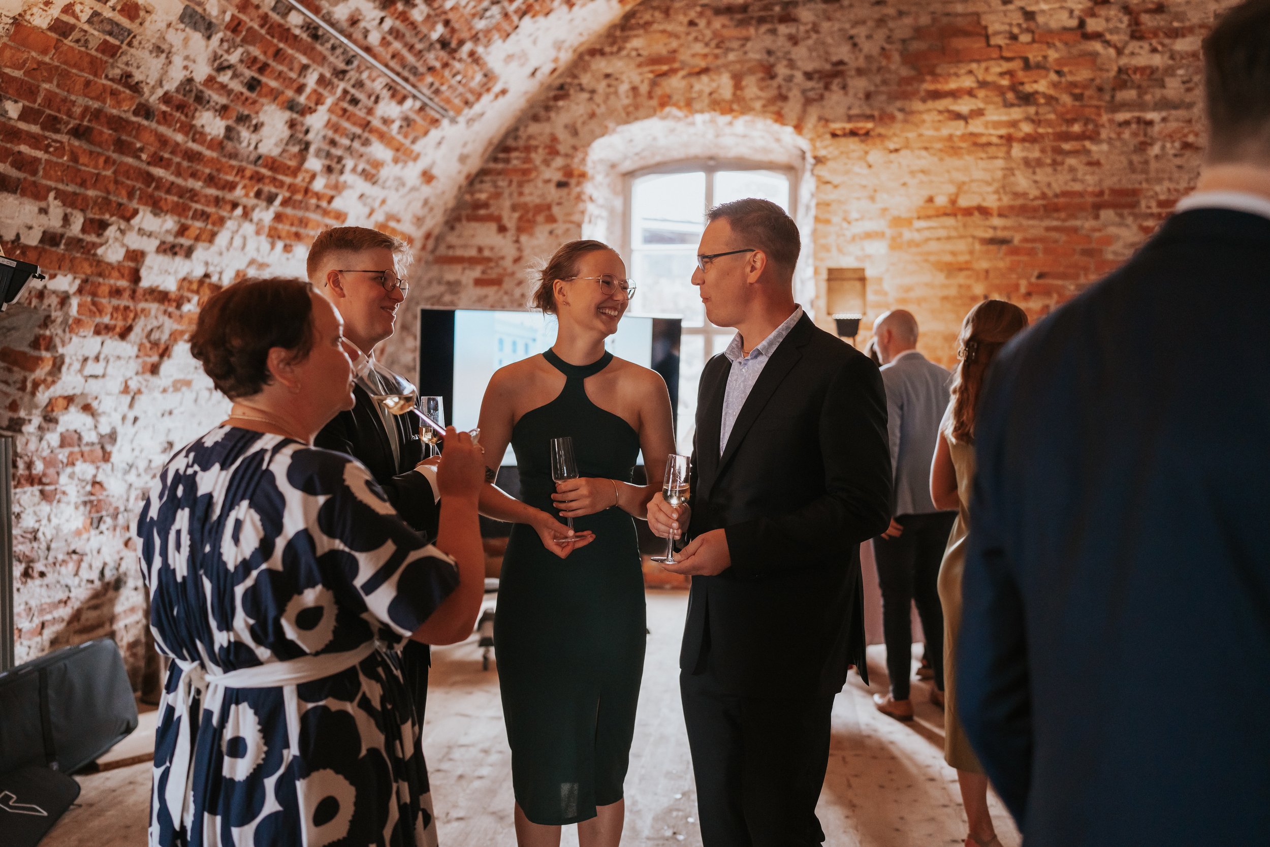 Group of people at an indoor event, smiling and holding glasses, against a rustic brick wall backdrop.