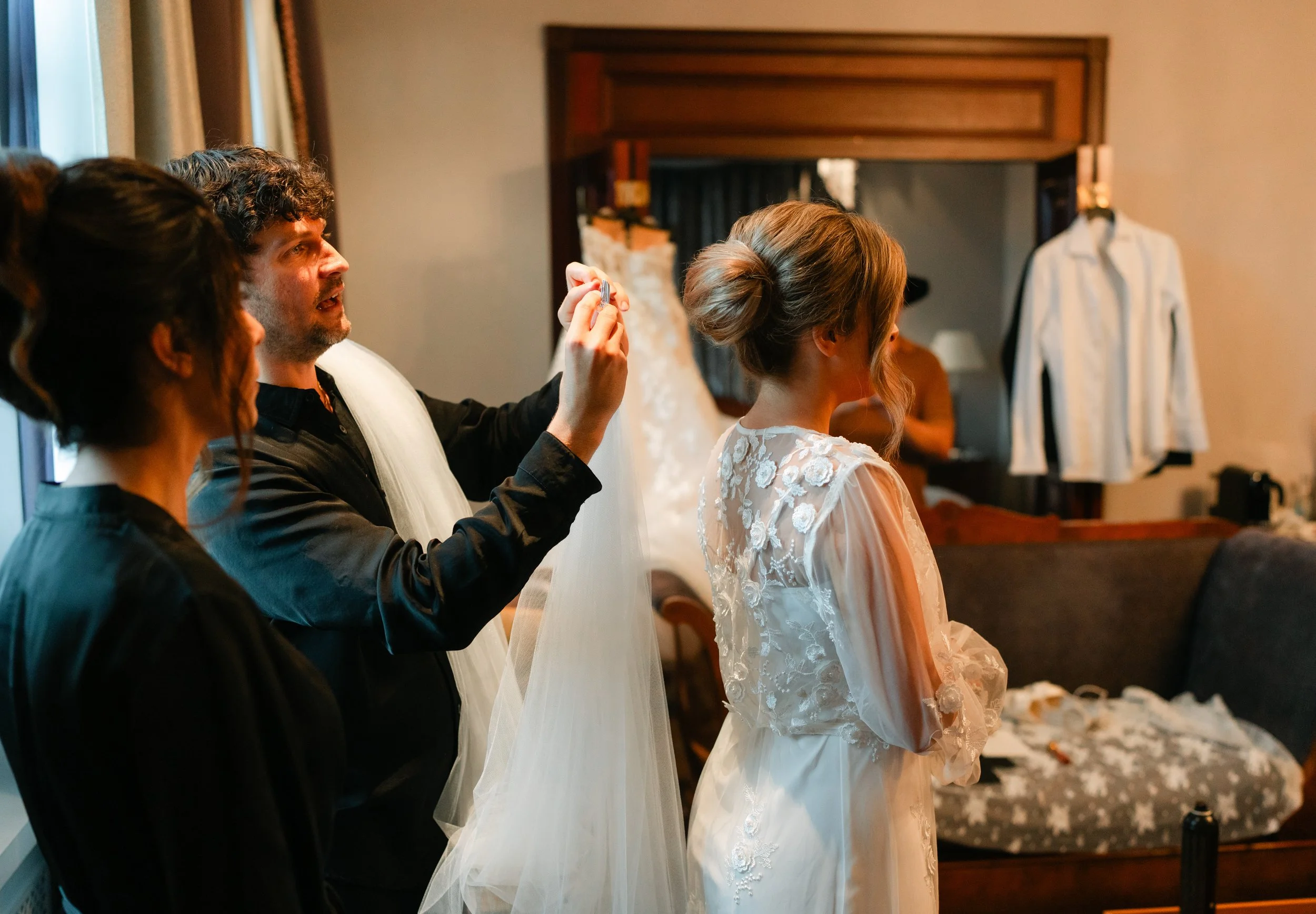 Bridal preparation scene with a stylist adjusting a veil on a bride wearing a lace wedding dress, surrounded by attendants in a room with a hanging shirt and sofa.