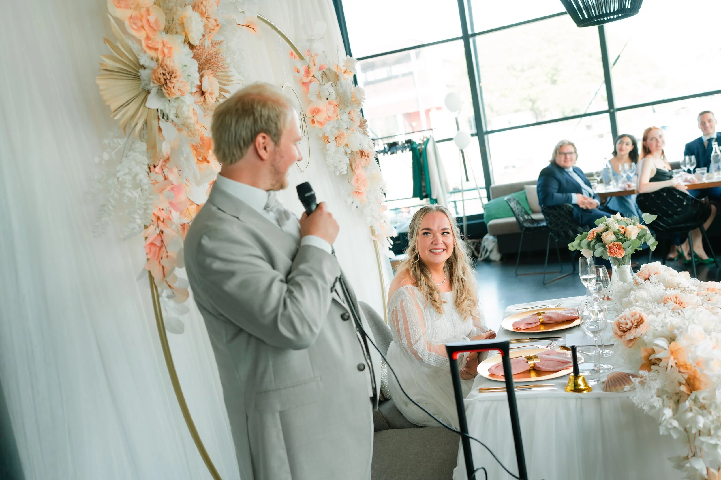 Wedding scene with a man in a suit speaking into a microphone and a woman in a wedding dress sitting at a table decorated with flowers, surrounded by guests in a bright, modern venue.