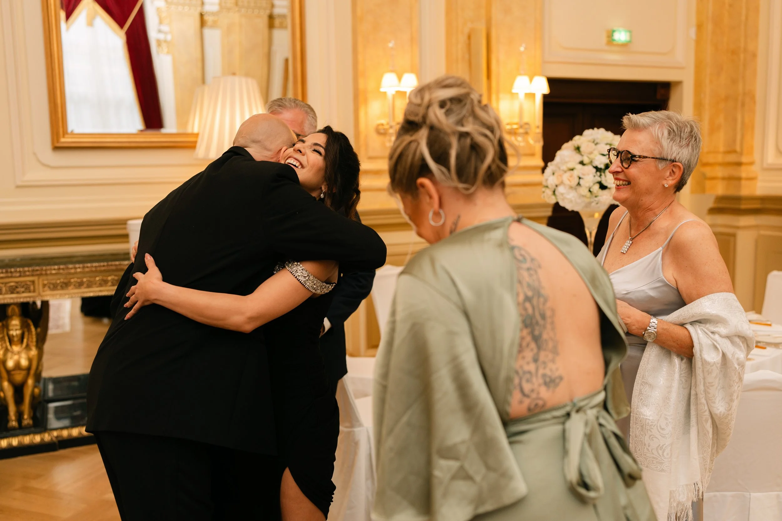 People hugging and smiling at a formal event in an elegant room with ornate decor.