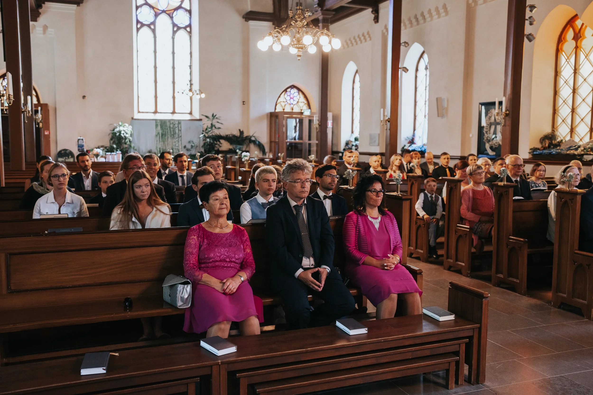 People sitting in a church during a ceremony, wearing formal attire, with stained glass windows and wooden pews visible.