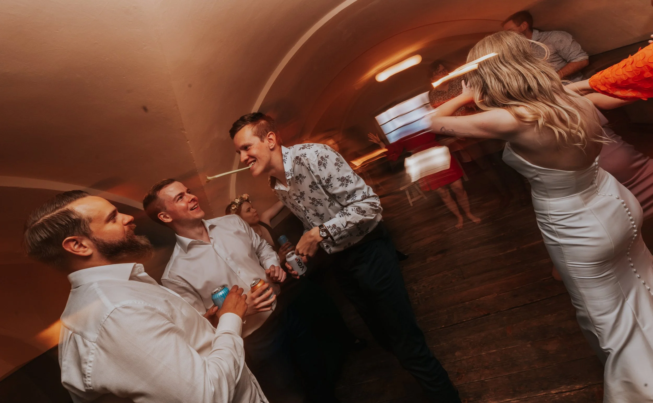 People dancing at a party indoors, wearing formal attire, with a low ceiling and wooden floor.