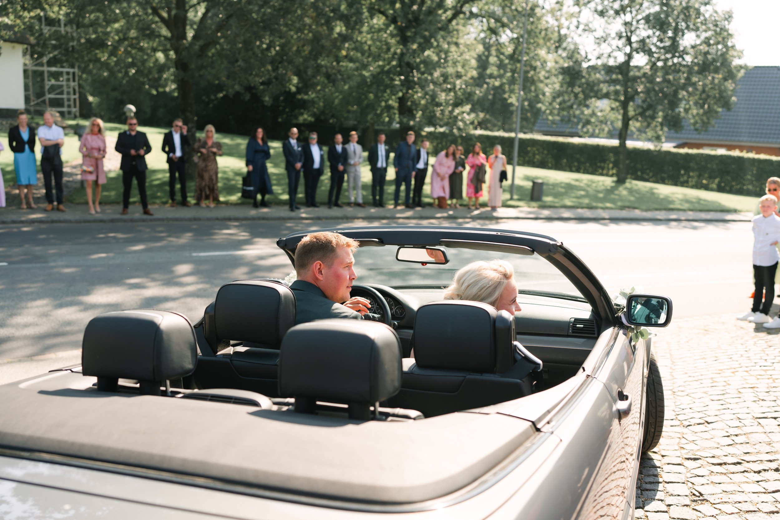 A man and woman in a convertible surrounded by people on the roadside, trees in the background.