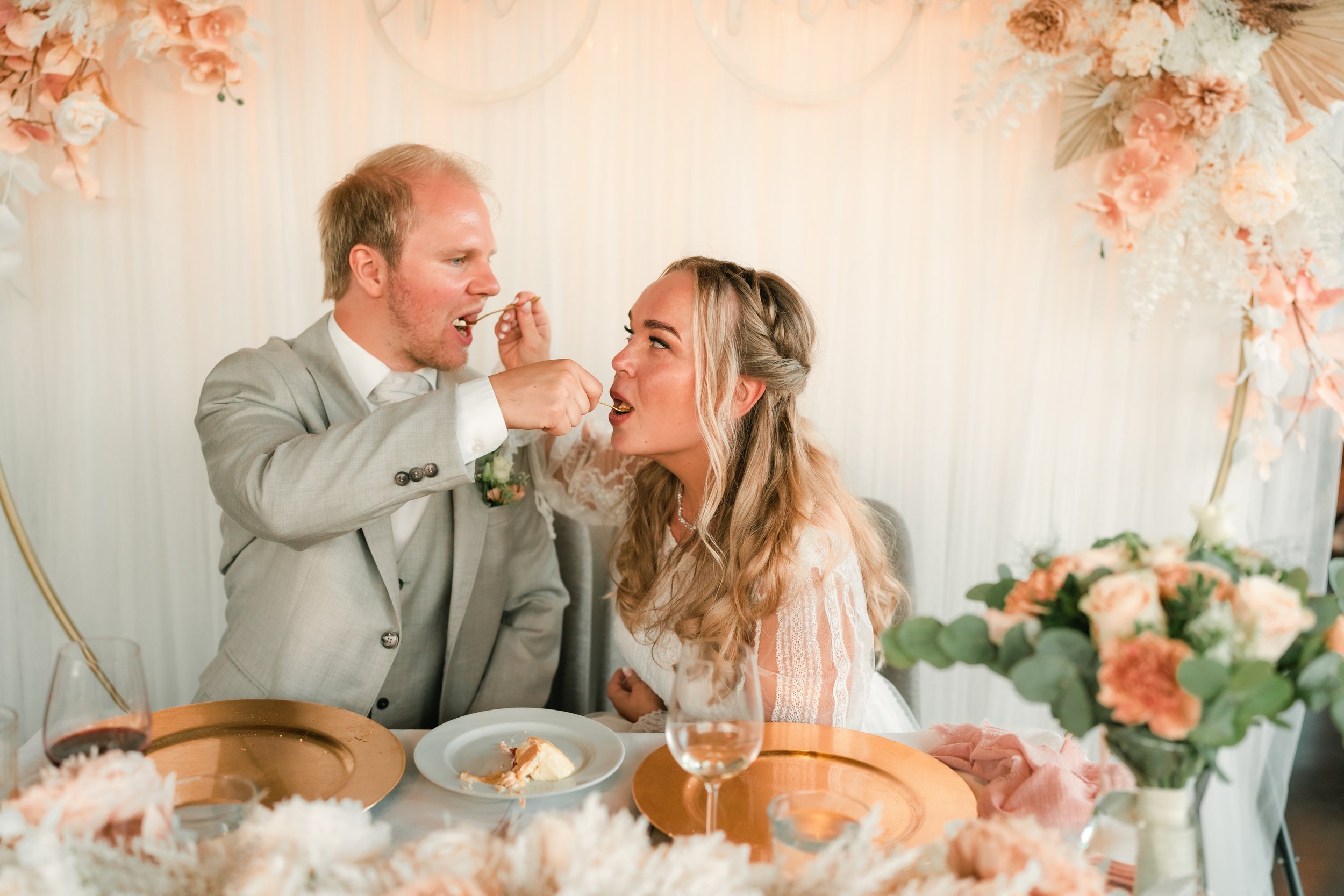 Bride and groom feeding each other cake at wedding reception, surrounded by elegant floral decorations and table setting.