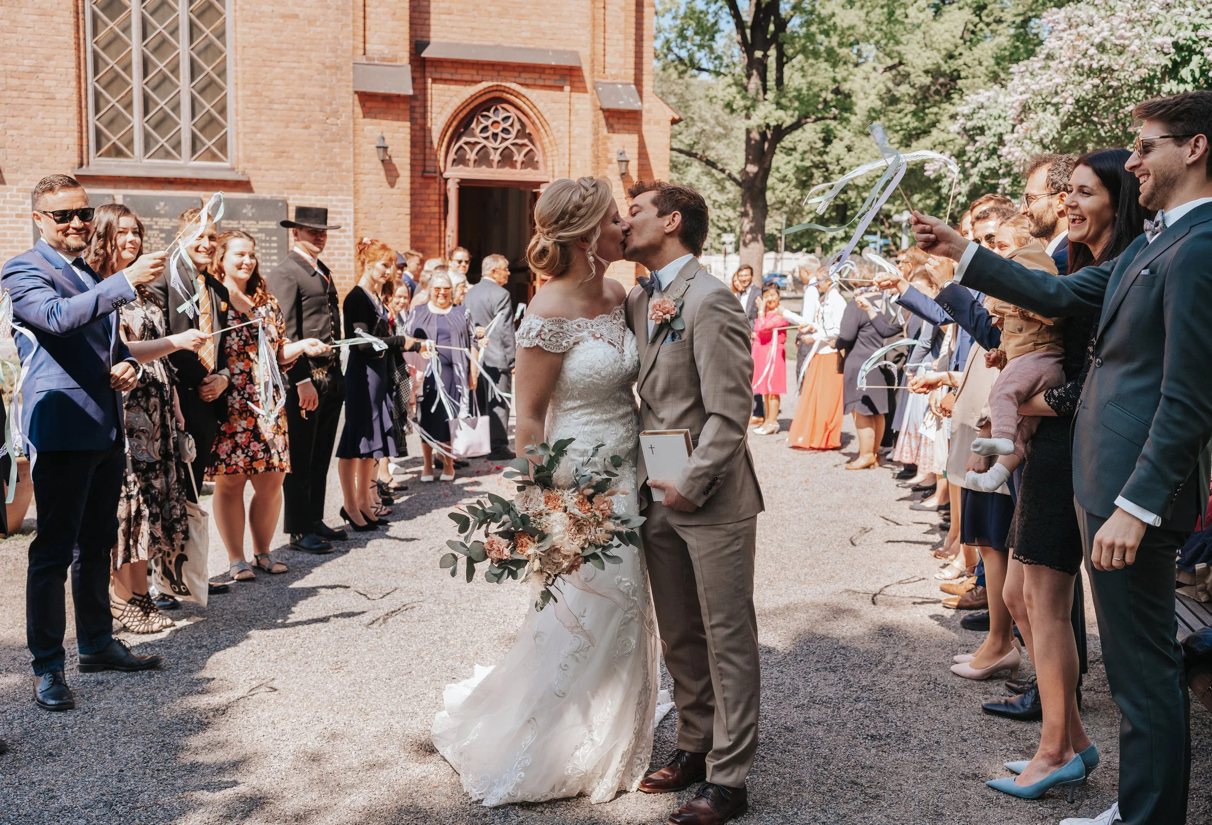 Bride and groom kissing outside a church surrounded by guests holding ribbon wands, wedding celebration.