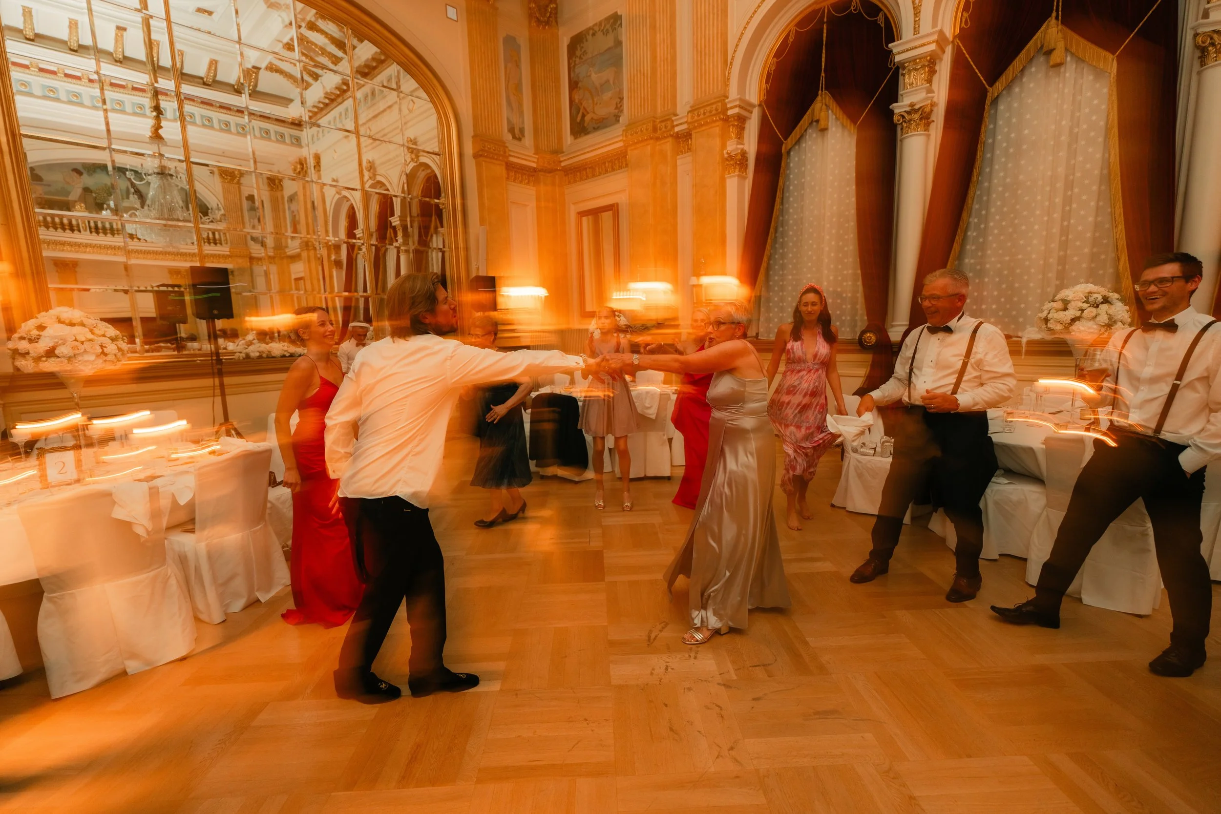 People dancing in a formal ballroom with ornate decor and large mirrors, capturing a lively party atmosphere.