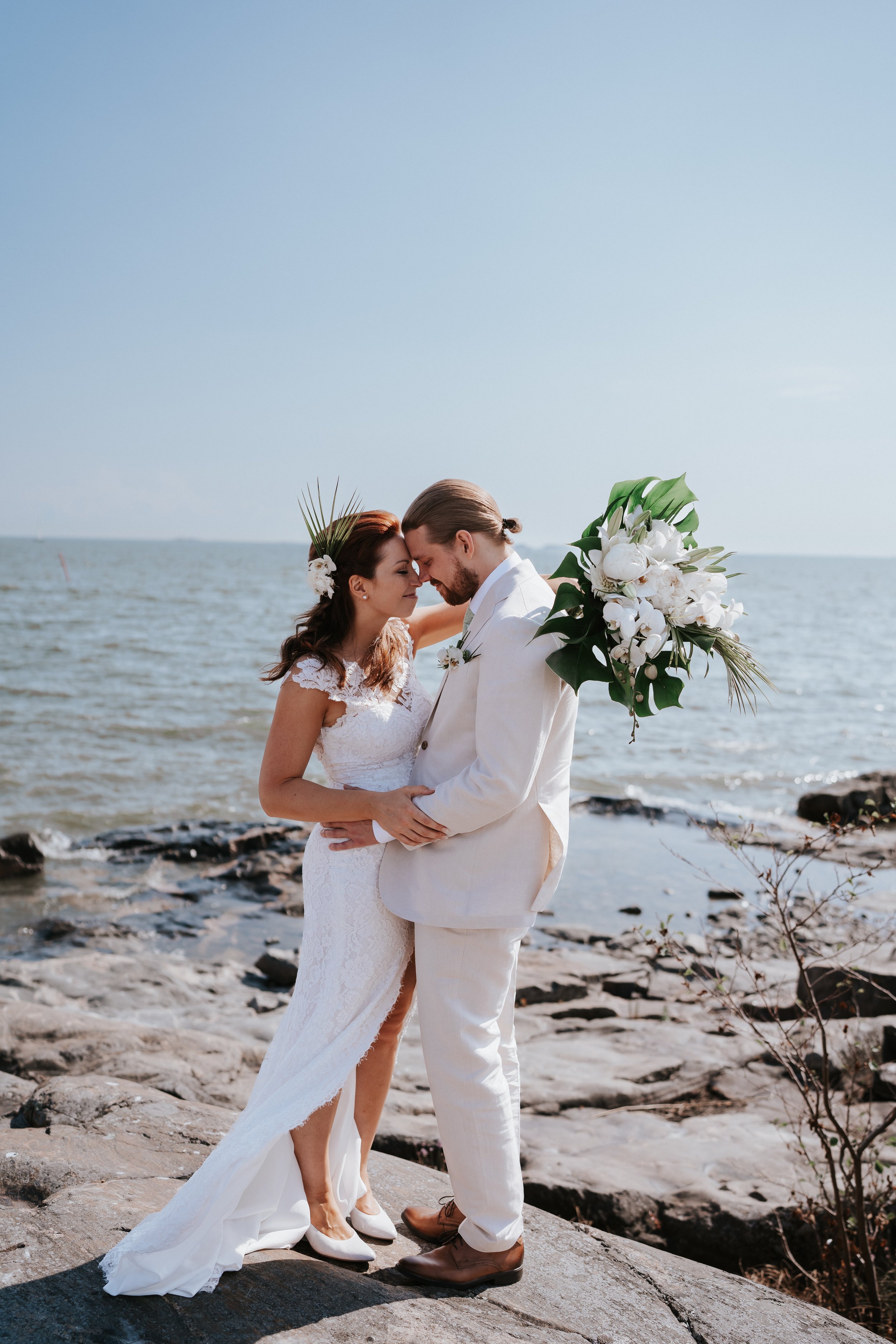 Bride and groom in white embracing on rocky shore by the ocean, holding bouquet of white flowers.