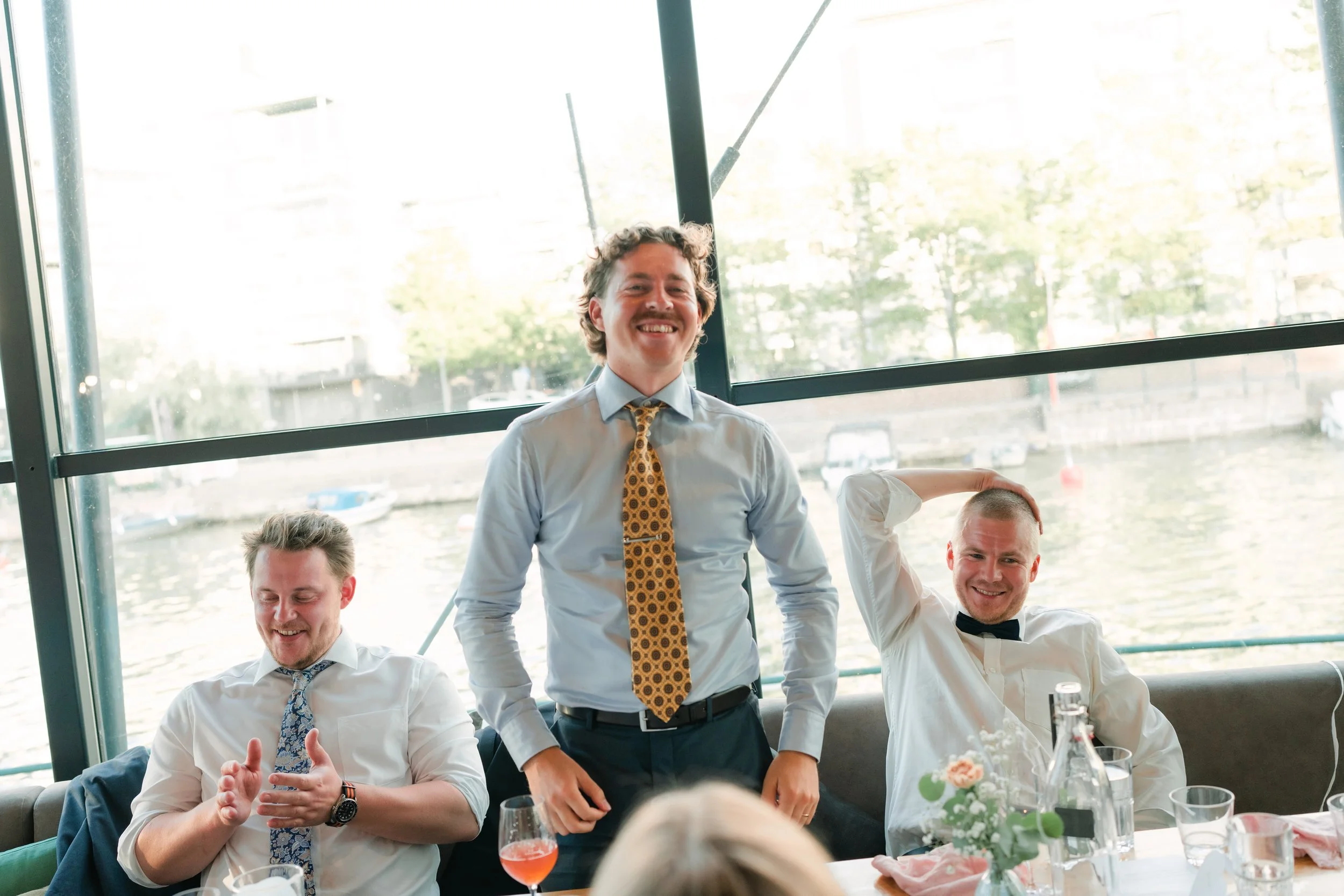 Three men enjoying a lighthearted moment indoors, with a water view in the background. One man is standing and smiling, wearing a light blue shirt and a patterned tie, while the other two are seated, appearing relaxed and happy.