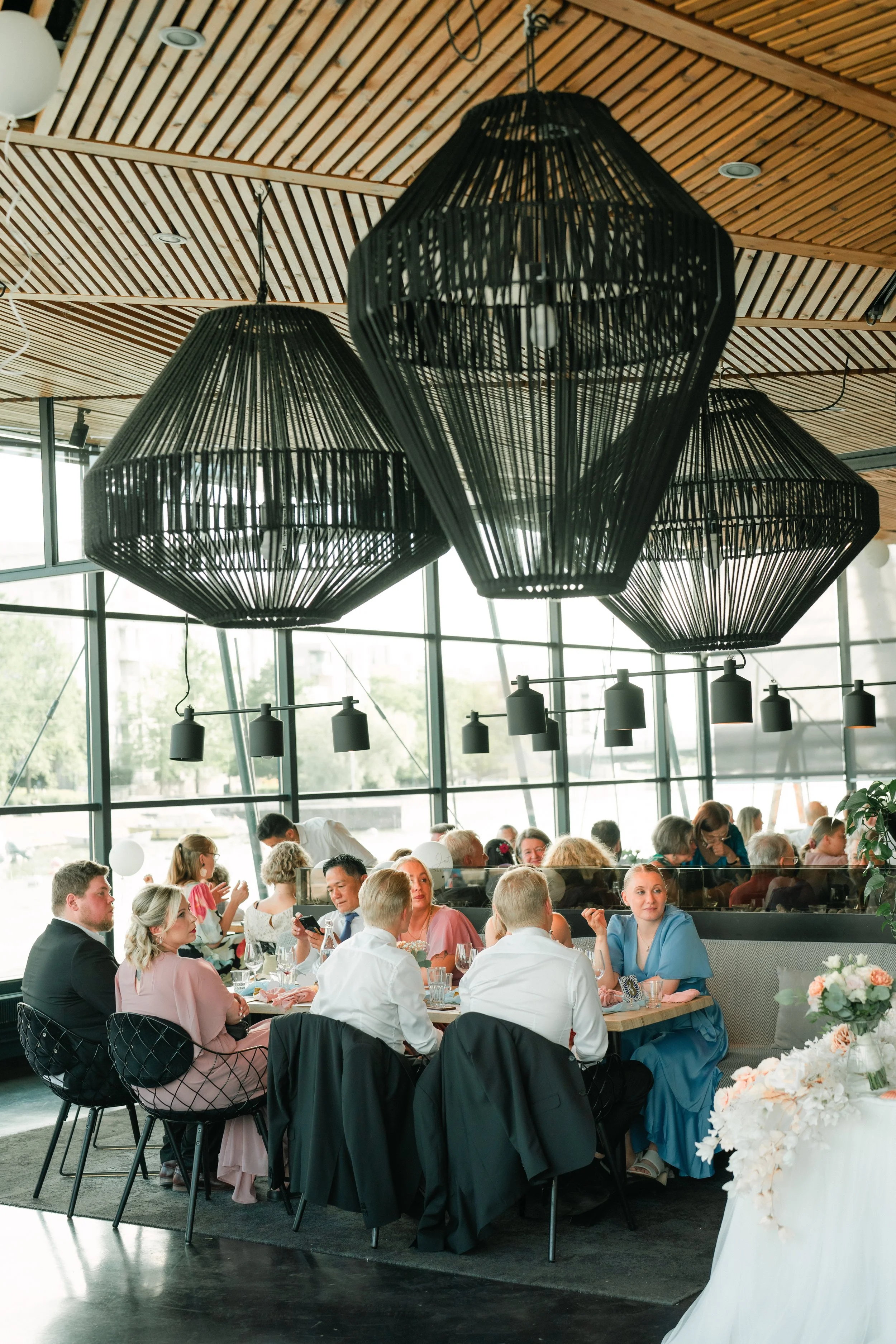 Elegant dining event with large black geometric pendant lights, modern interior design, guests seated at a long table, floral decorations.