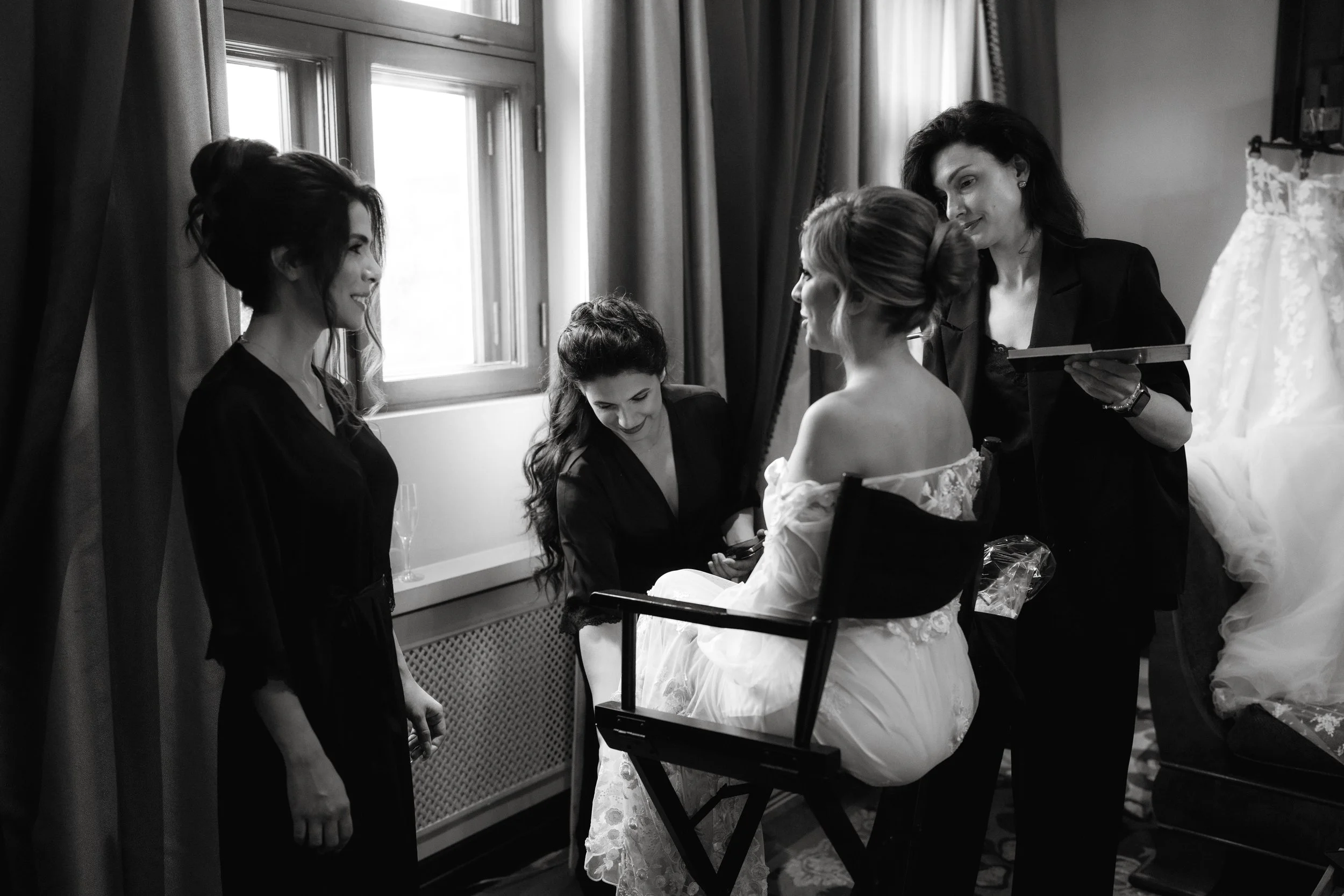 Bridal party getting ready, bride in makeup chair, black and white photo, window, curtains, three stylists assisting, wedding dress in background.