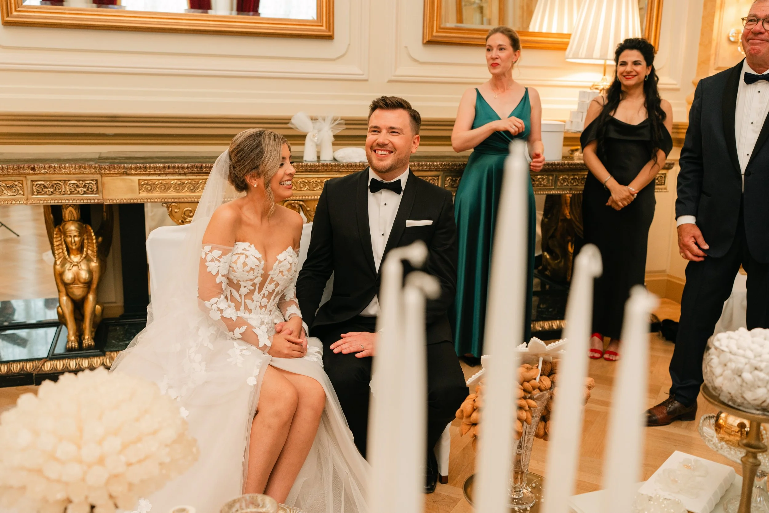 Bride and groom sitting at a wedding ceremony with guests, candles, and decorative items.